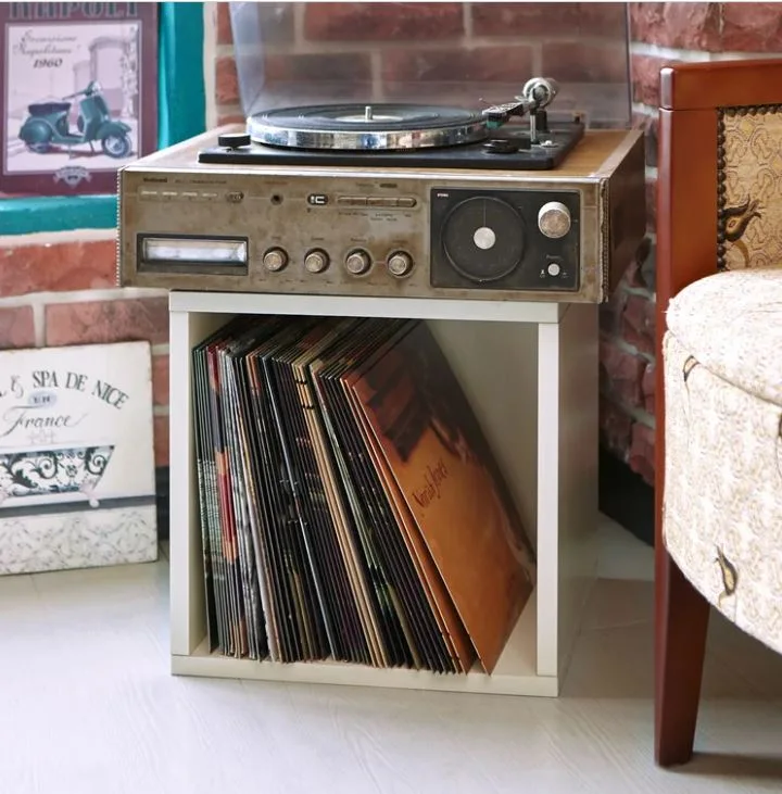 A vintage record player sits on a white storage cube filled with vinyl records against an exposed red brick wall.