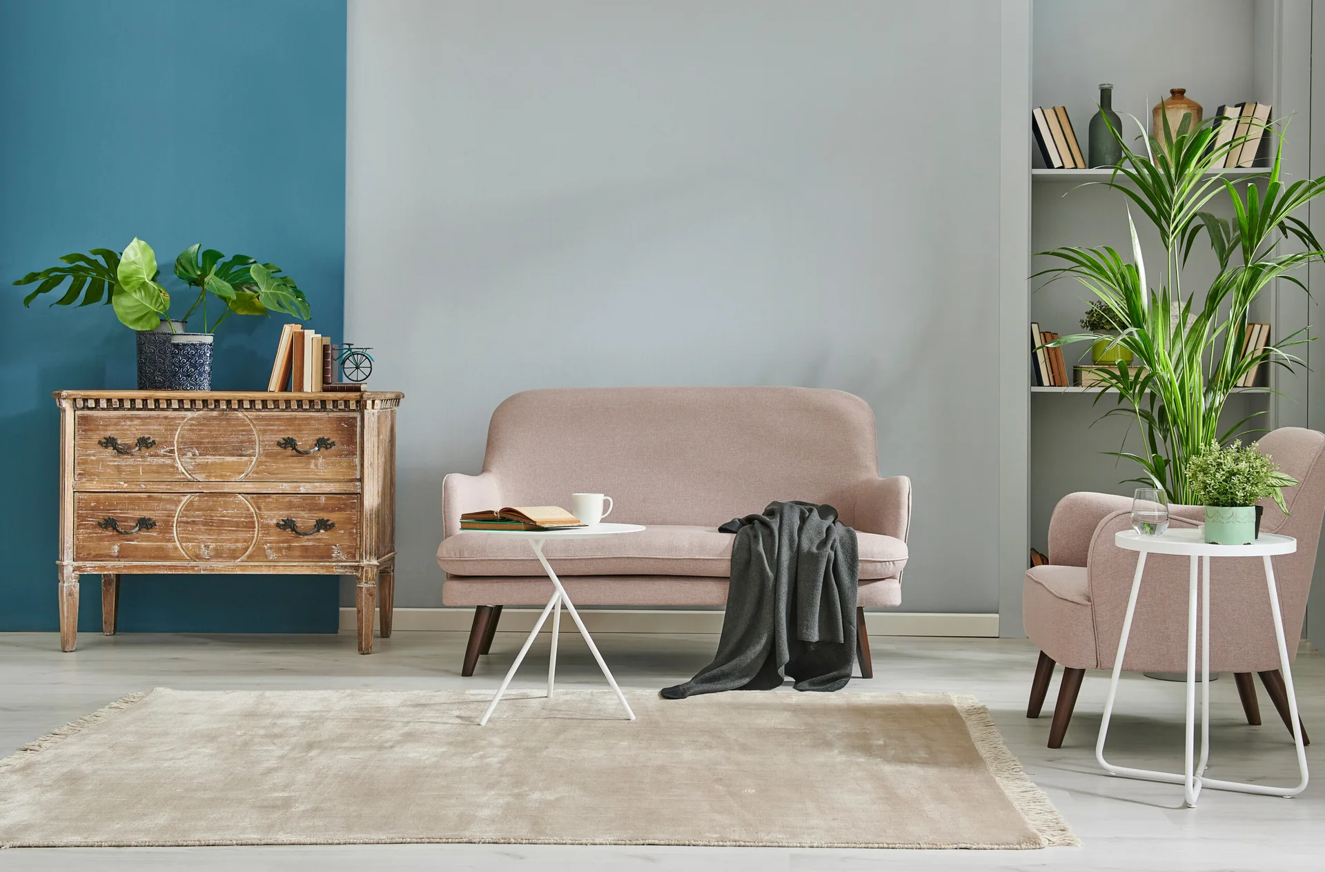 A living room featuring a blush pink sofa and armchair against a blue and grey painted wall, next to a distressed wood dresser and green houseplants.