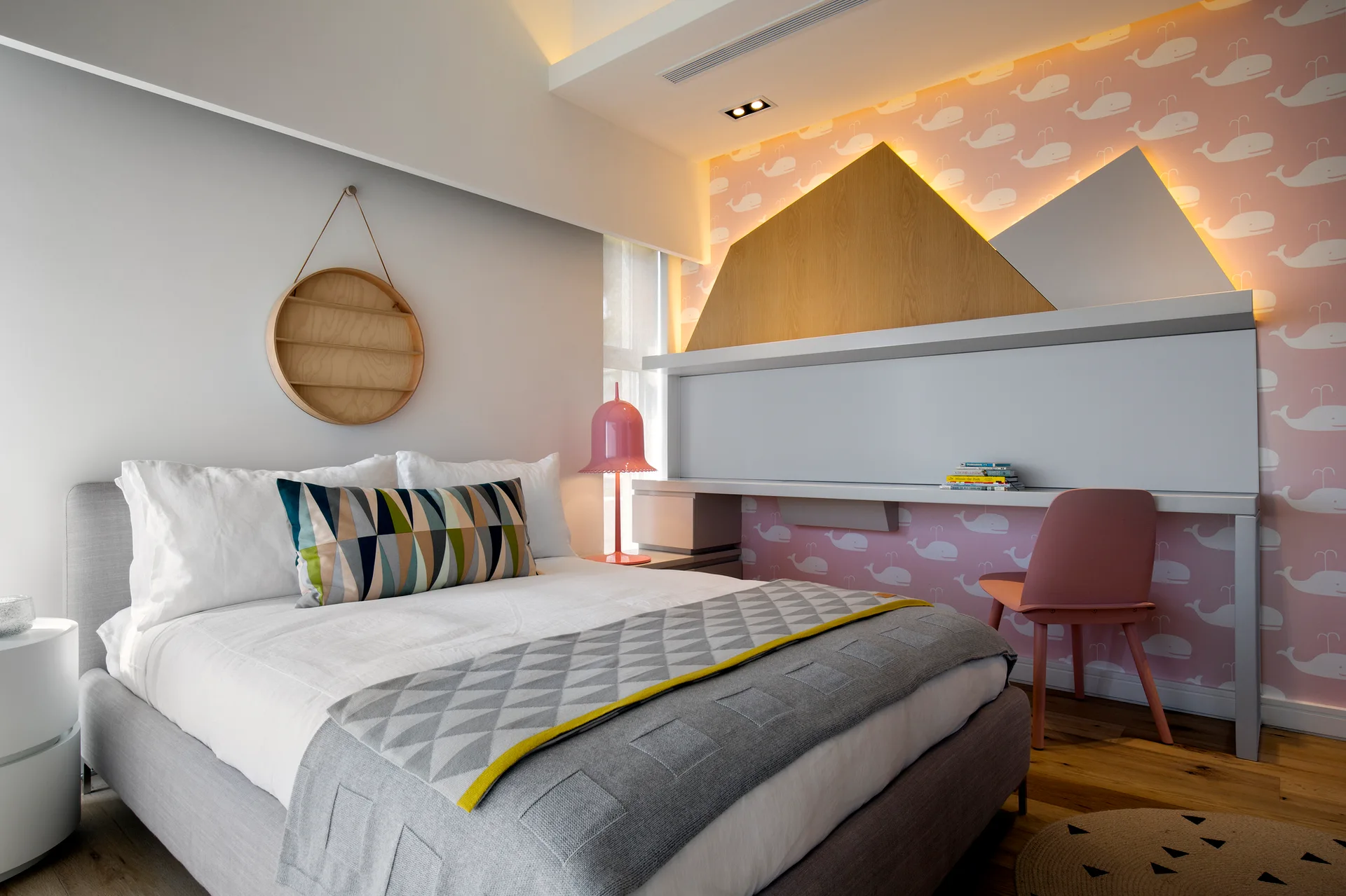 A kids bedroom featuring a gray upholstered bed, pink whale-patterned wallpaper, and an integrated study desk with mountain-shaped wall panels.