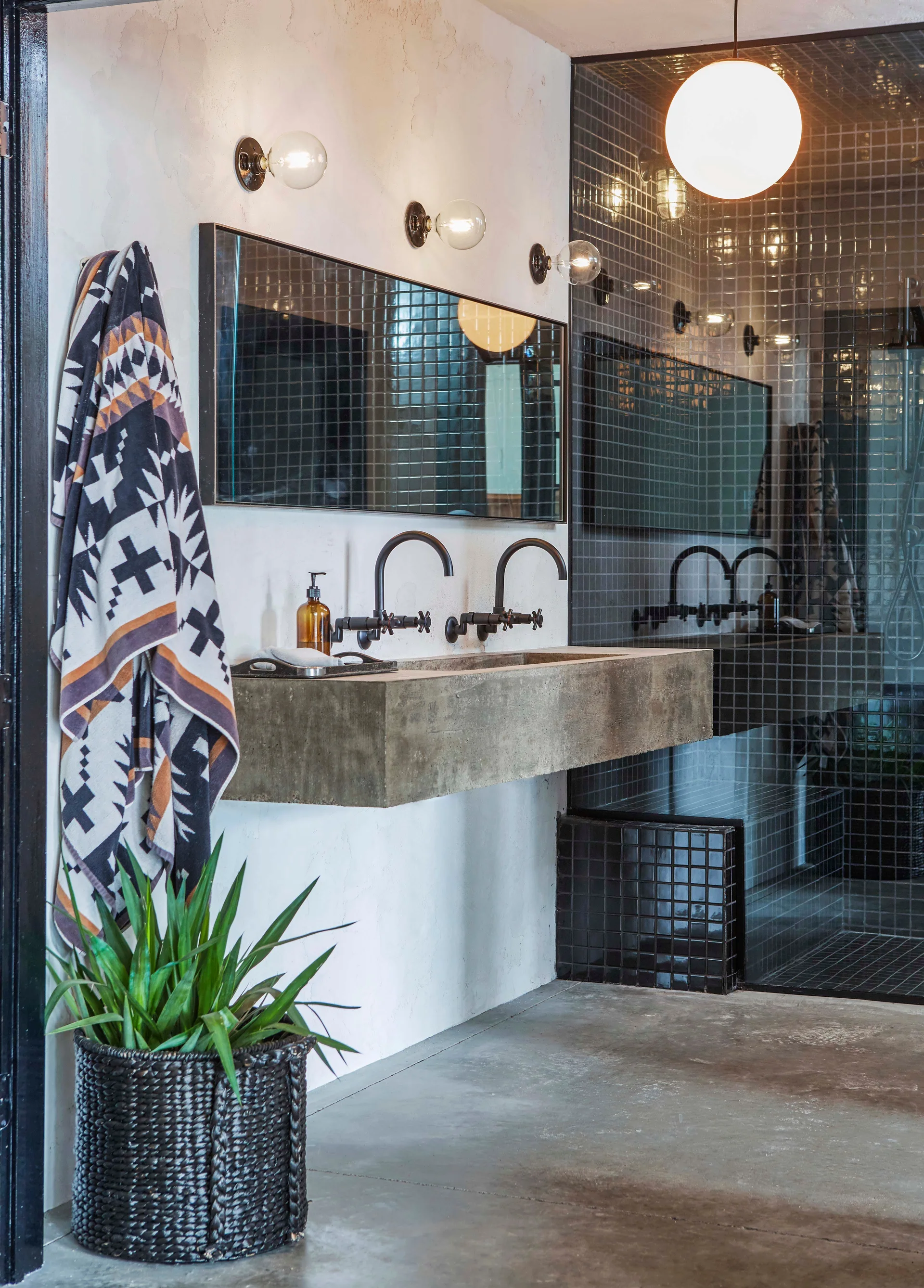 An industrial bathroom features a wall-mounted concrete trough sink with black faucets beneath a long mirror and black-tiled shower.