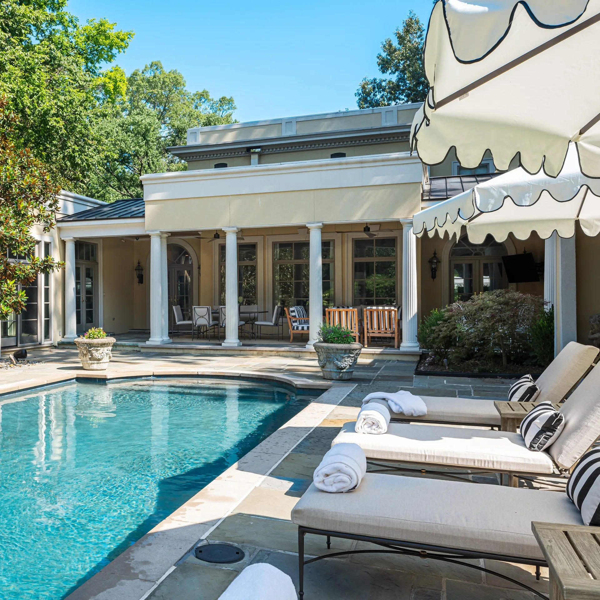 A backyard swimming pool is lined with tan lounge chairs, striped pillows, and scalloped umbrellas next to a stone patio with white columns.