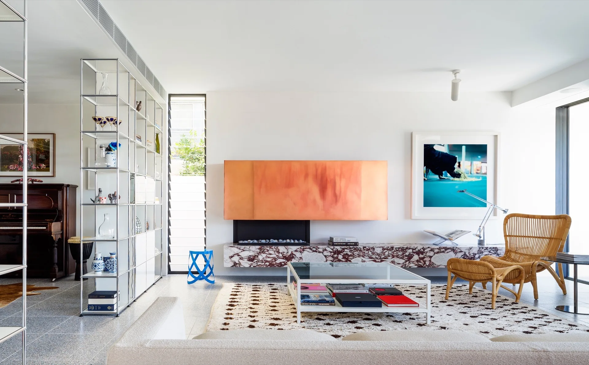 A contemporary living room features a striking copper-paneled fireplace, a low marble hearth, a rattan lounge chair, and white modular shelving.