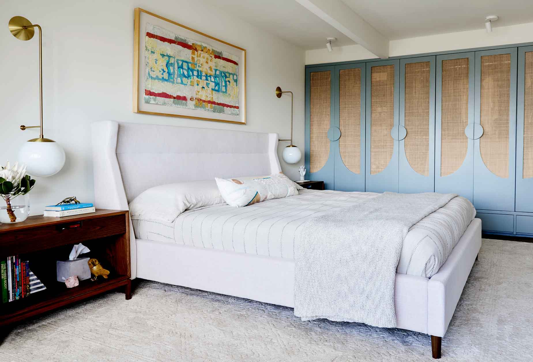A modern bedroom features an upholstered white bed next to a dark wood nightstand and a large built-in blue wardrobe with natural cane panels.