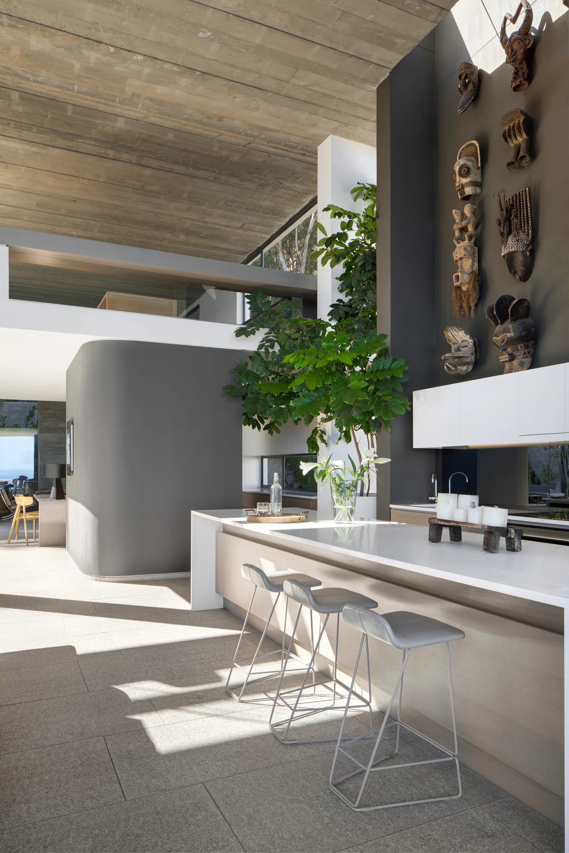 A modern kitchen features a light wood island with grey stools, an exposed concrete ceiling, and a tall charcoal accent wall decorated with carved wooden masks.