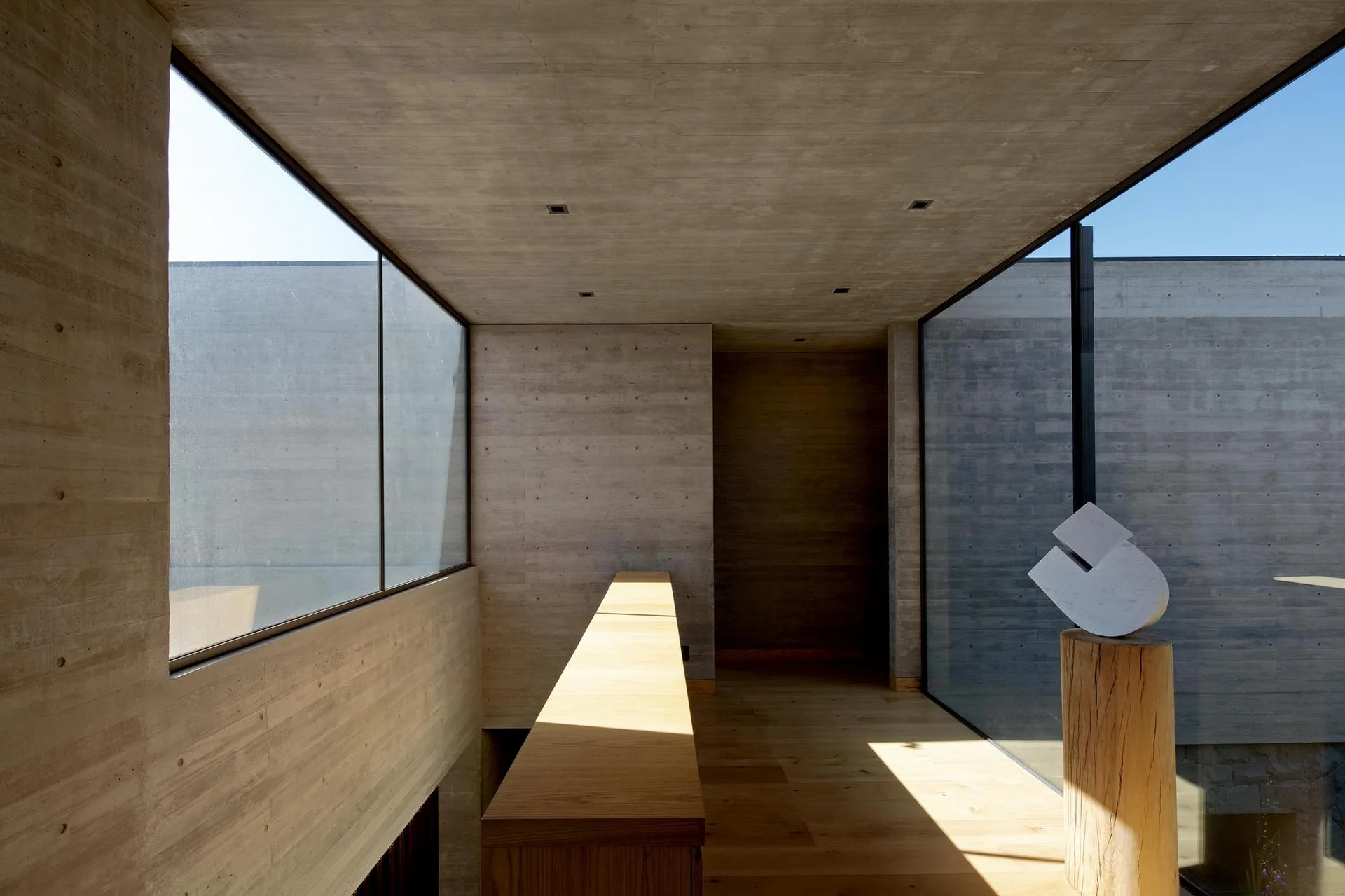 A minimalist hallway features board-formed concrete walls, light wood plank floors, a long timber console, and a large window looking outside.