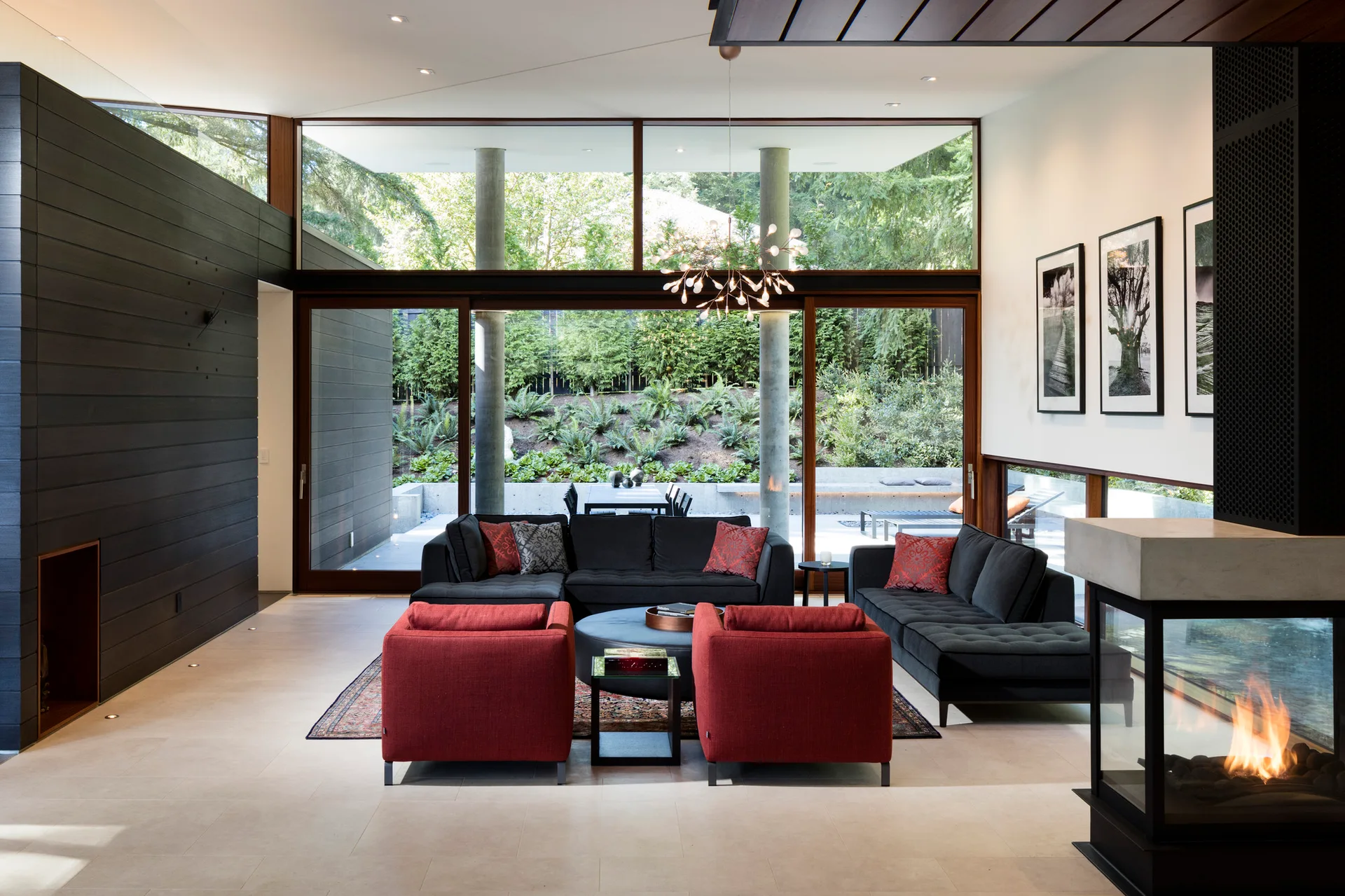A contemporary living room featuring a black sectional sofa, two red armchairs, a glass fireplace, and floor-to-ceiling windows showing a garden.
