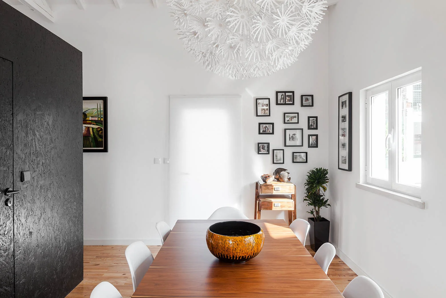 A modern dining room featuring a long wooden table, white plastic chairs, an oversized spiky pendant light, and a textured black accent wall.