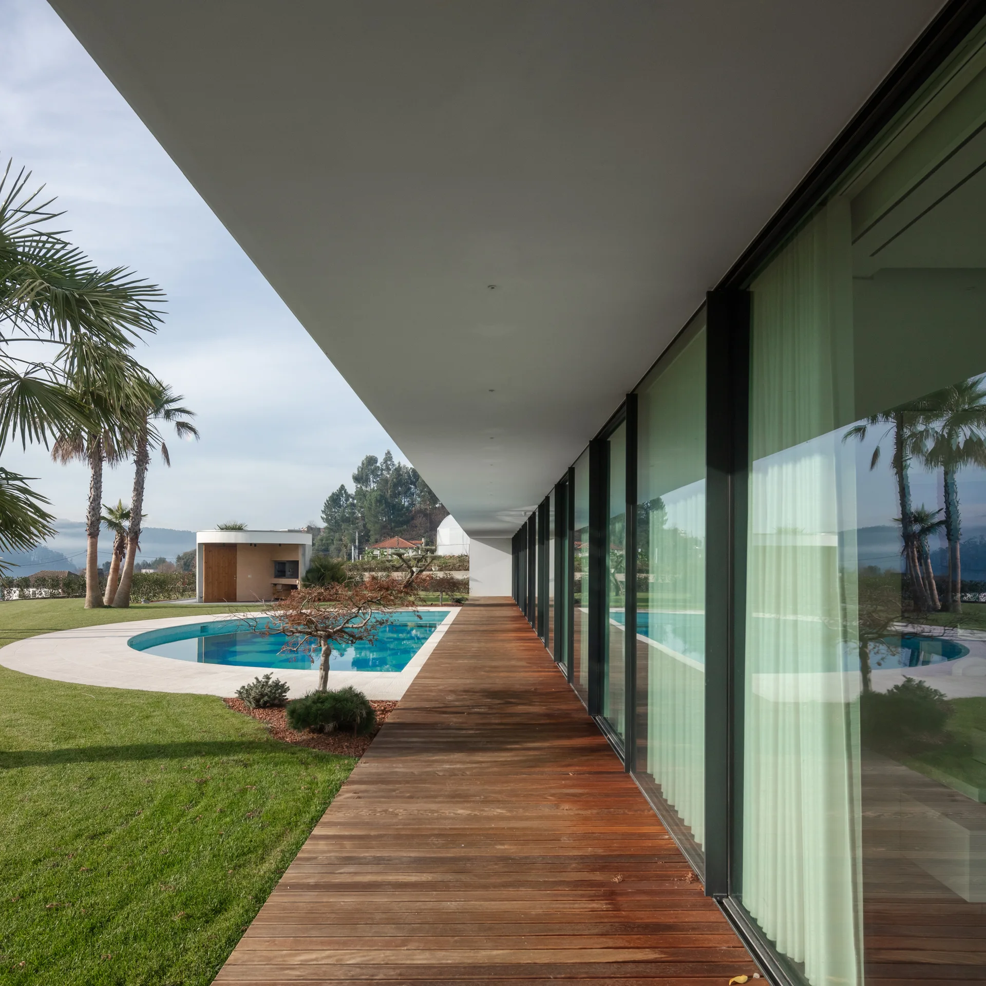 A modern home features a long wooden plank deck running alongside a floor-to-ceiling glass facade and a curved outdoor swimming pool.