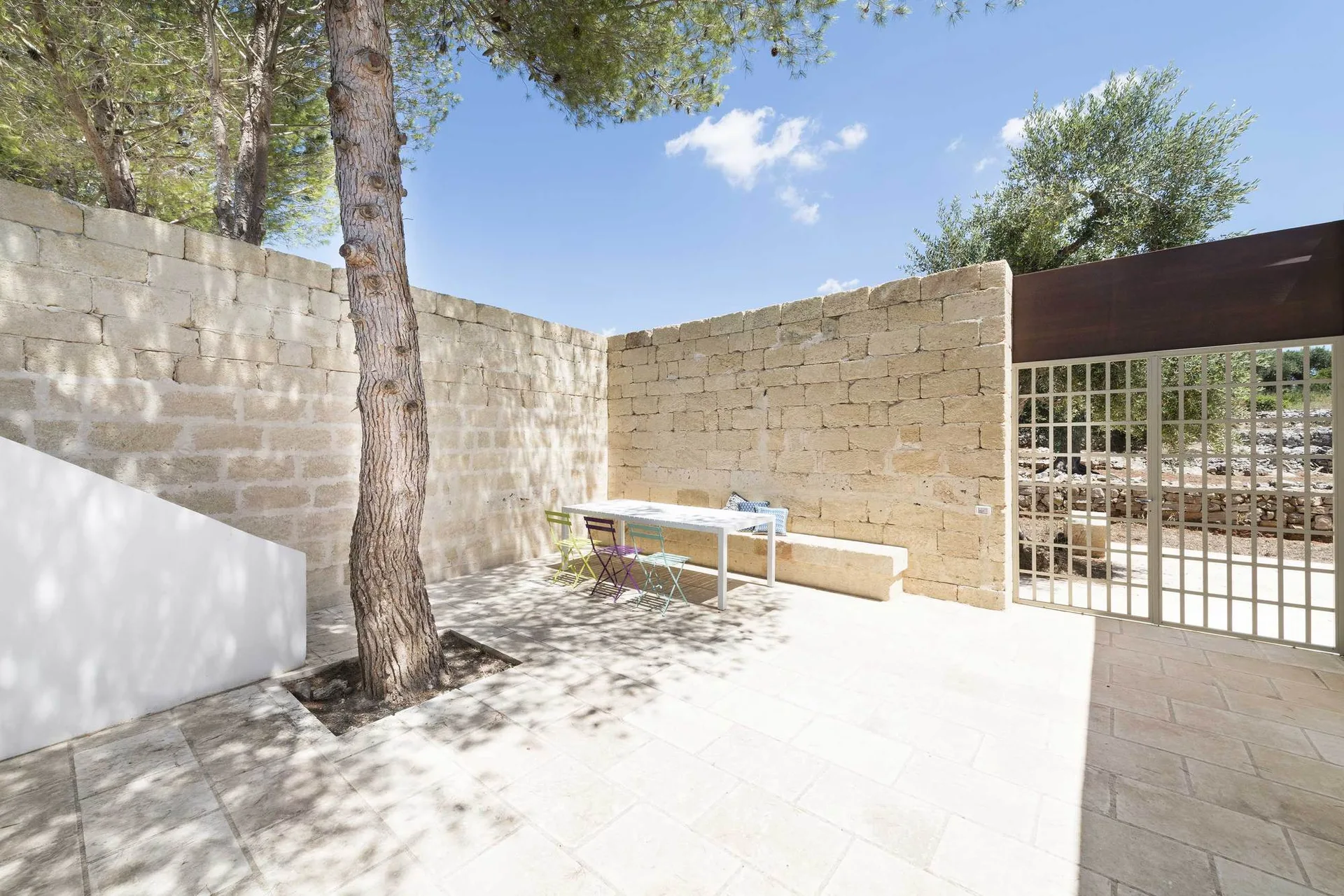 An outdoor stone patio features a white dining table with colorful folding chairs next to a built-in stone bench and a mature tree.