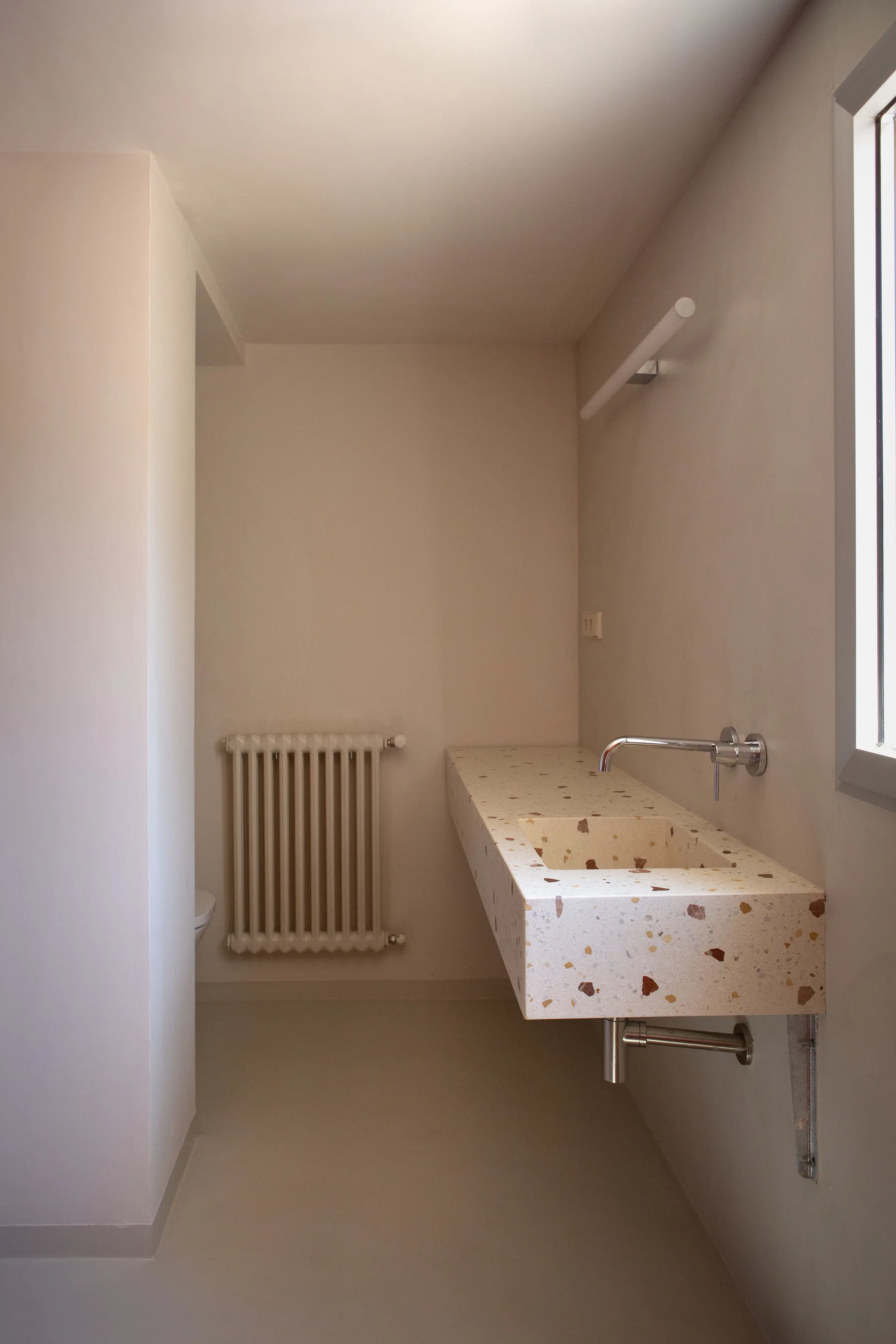 A minimalist cream bathroom featuring a wall-mounted speckled terrazzo vanity, chrome wall faucet, and a white classic radiator against the far wall.