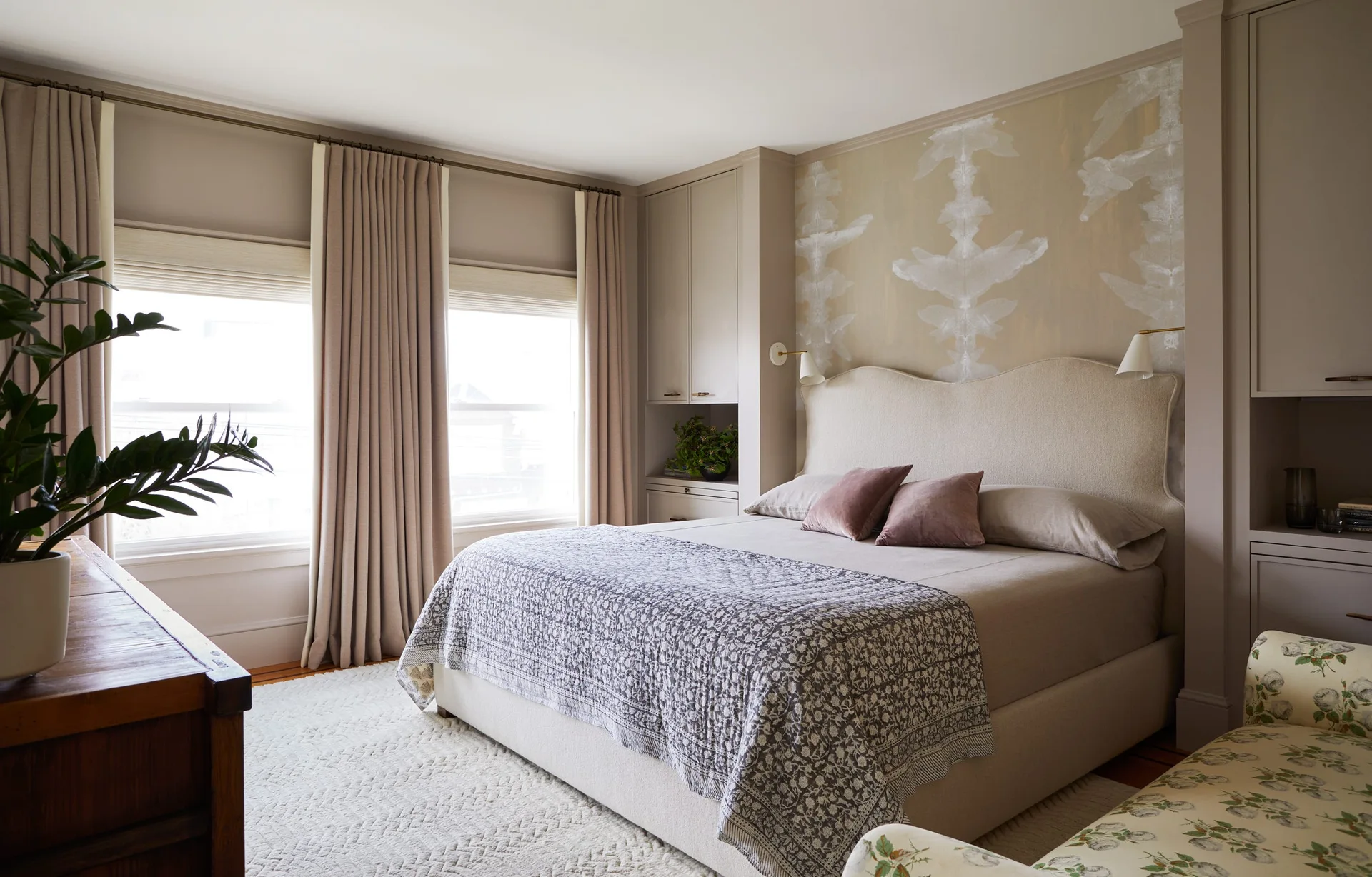A neutral bedroom features an upholstered bed with a scalloped headboard, flanked by custom taupe wardrobes and soft floor-length drapes.