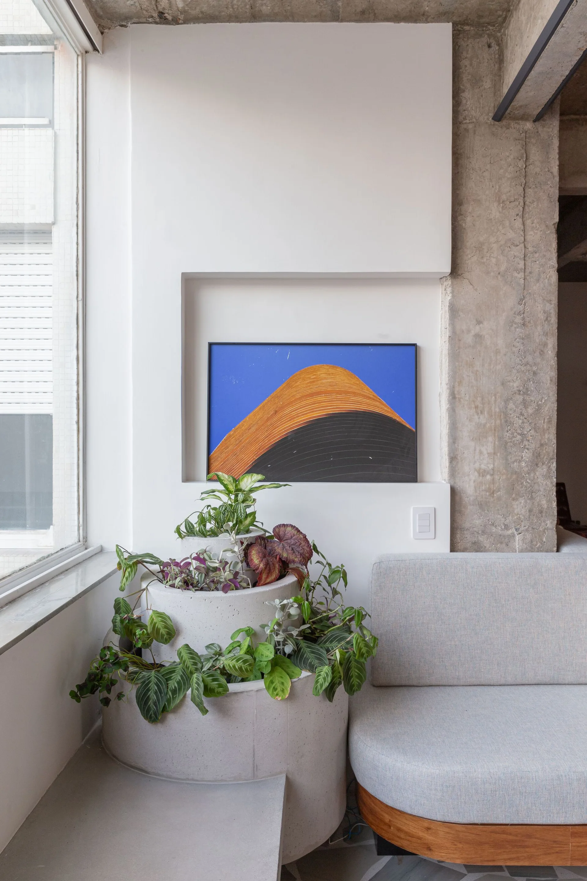 A light gray sofa sits beside a tiered concrete planter and a framed blue-and-orange abstract artwork set in a recessed wall niche.