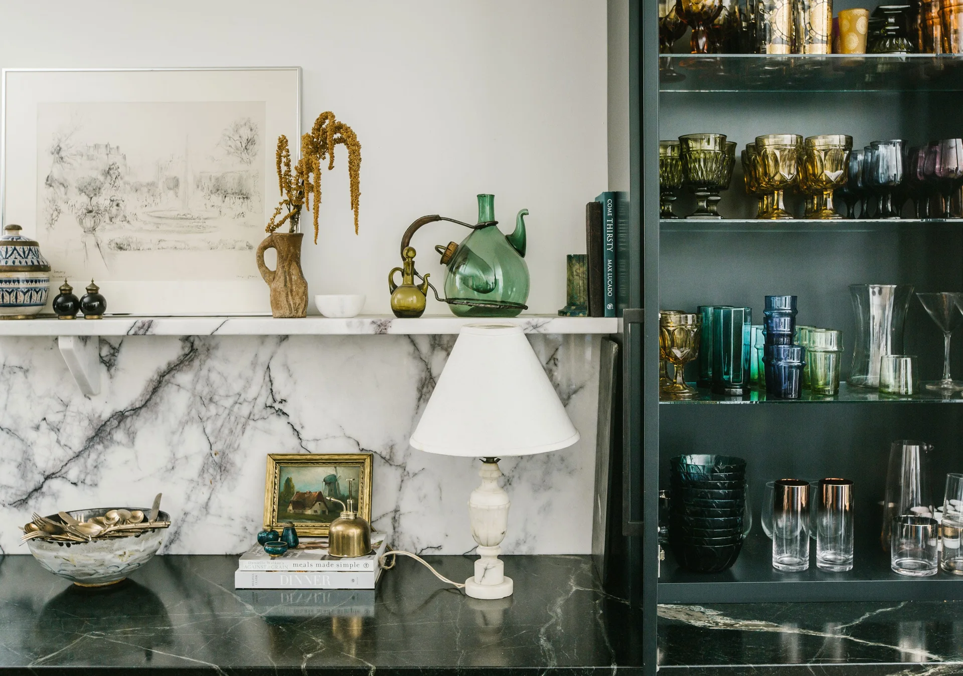 A kitchen counter area with a black marble countertop, white marble backsplash with a shelf, a table lamp, and a cabinet displaying colorful glassware.