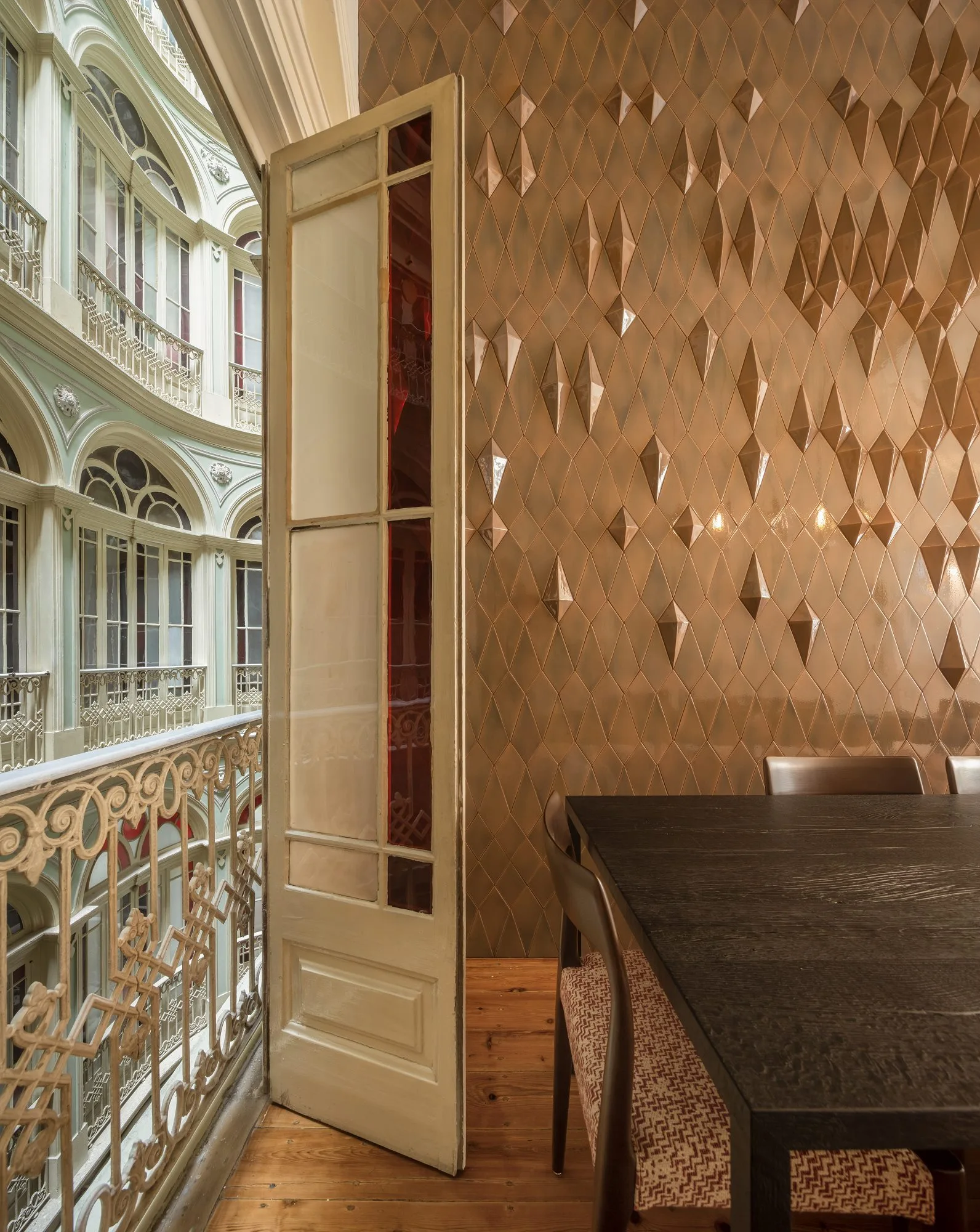 A dining room features a dark wood table and chairs next to a glossy bronze diamond-patterned textured wall and an open tall panel door leading to a balcony.