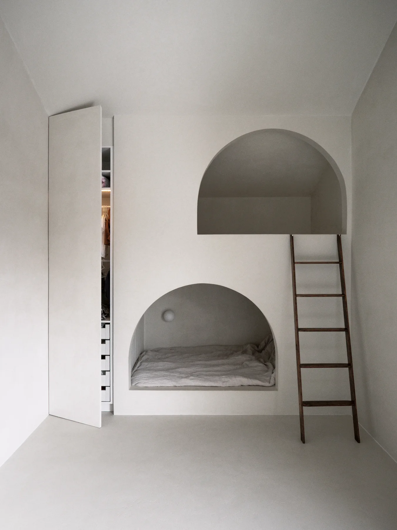 A minimalist bedroom features a built-in plaster wall structure with stacked arched sleeping nooks, a wooden ladder, and an open closet.