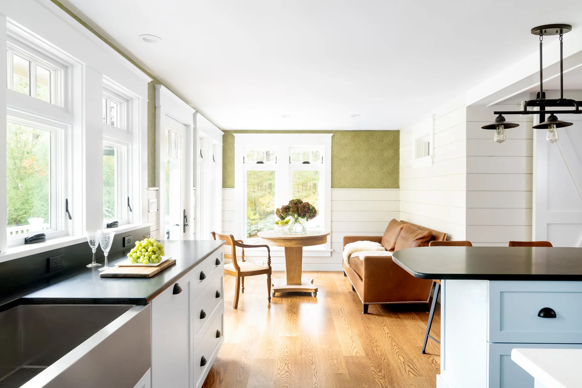 A bright farmhouse kitchen featuring white cabinetry, a leather sofa, and a round wooden pedestal table against shiplap and green wallpapered walls.