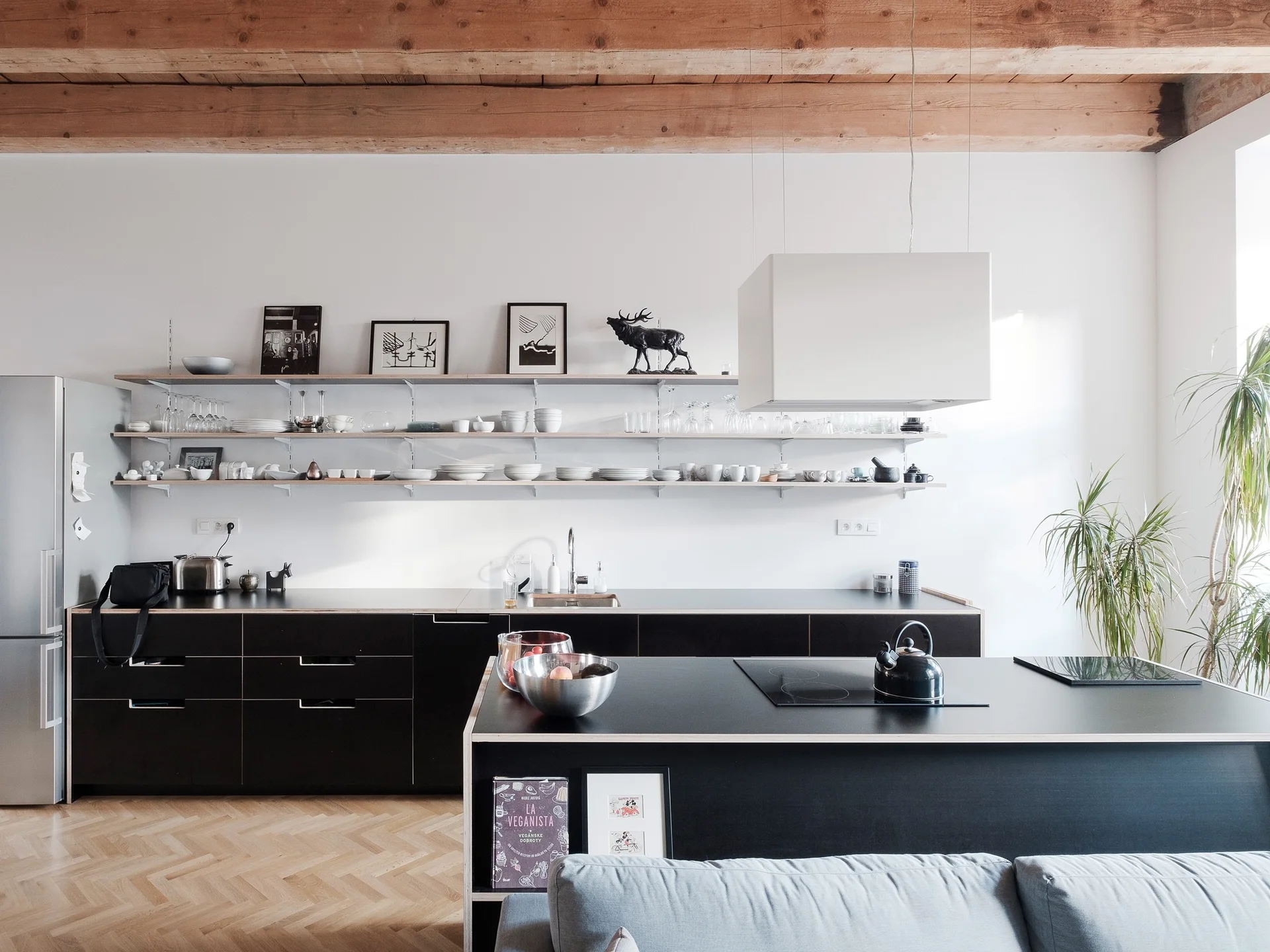 A modern kitchen featuring matte black cabinetry with plywood edges, long open shelves on a white wall, and exposed wooden ceiling beams.
