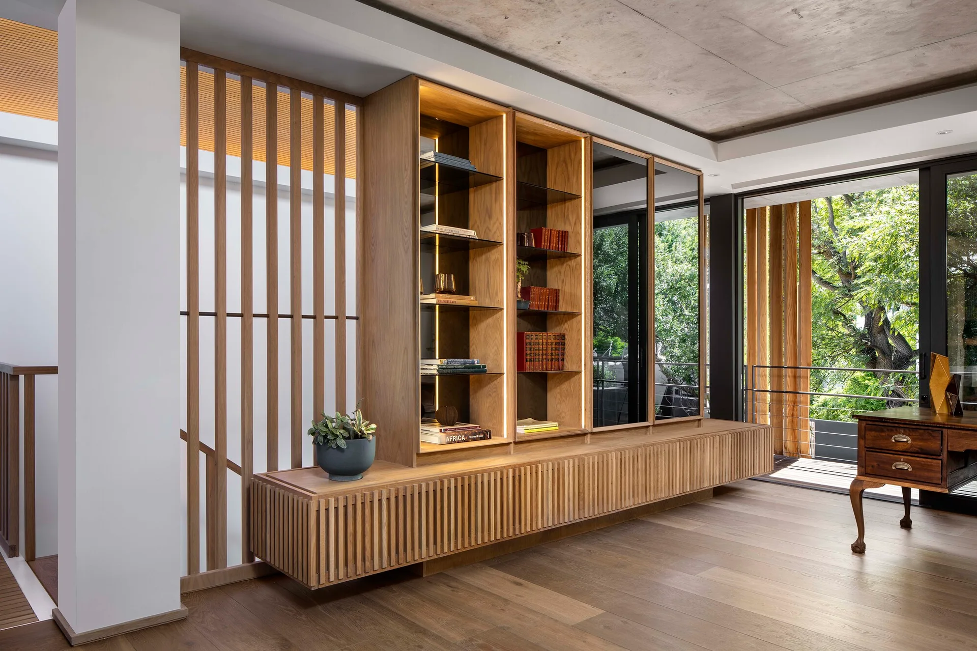 A custom oak shelving unit with integrated lighting stands next to a wooden slat room divider under a concrete ceiling.