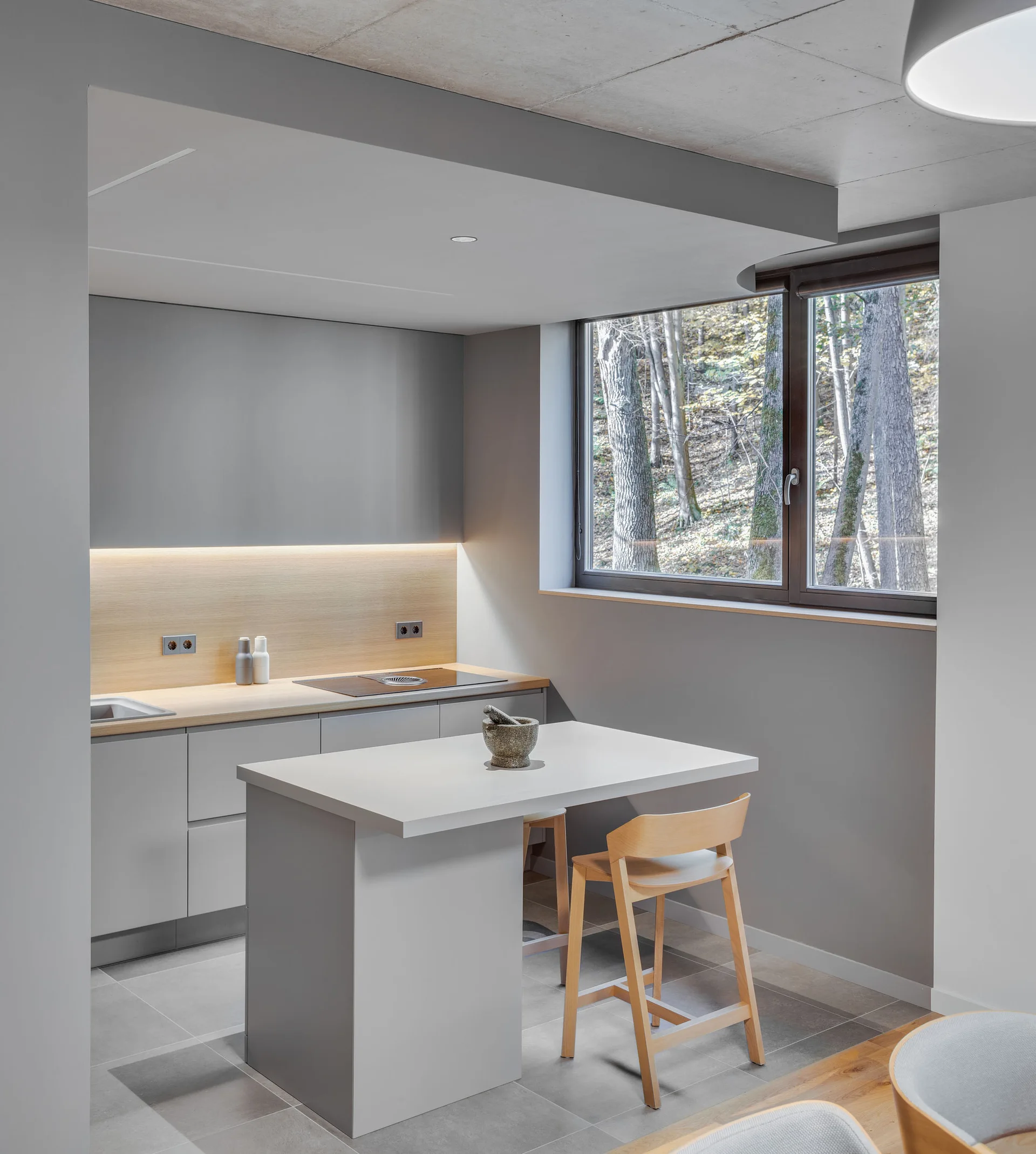A minimalist gray kitchen features a central island with wood stools, a matching wood backsplash, and a window displaying a forest view.