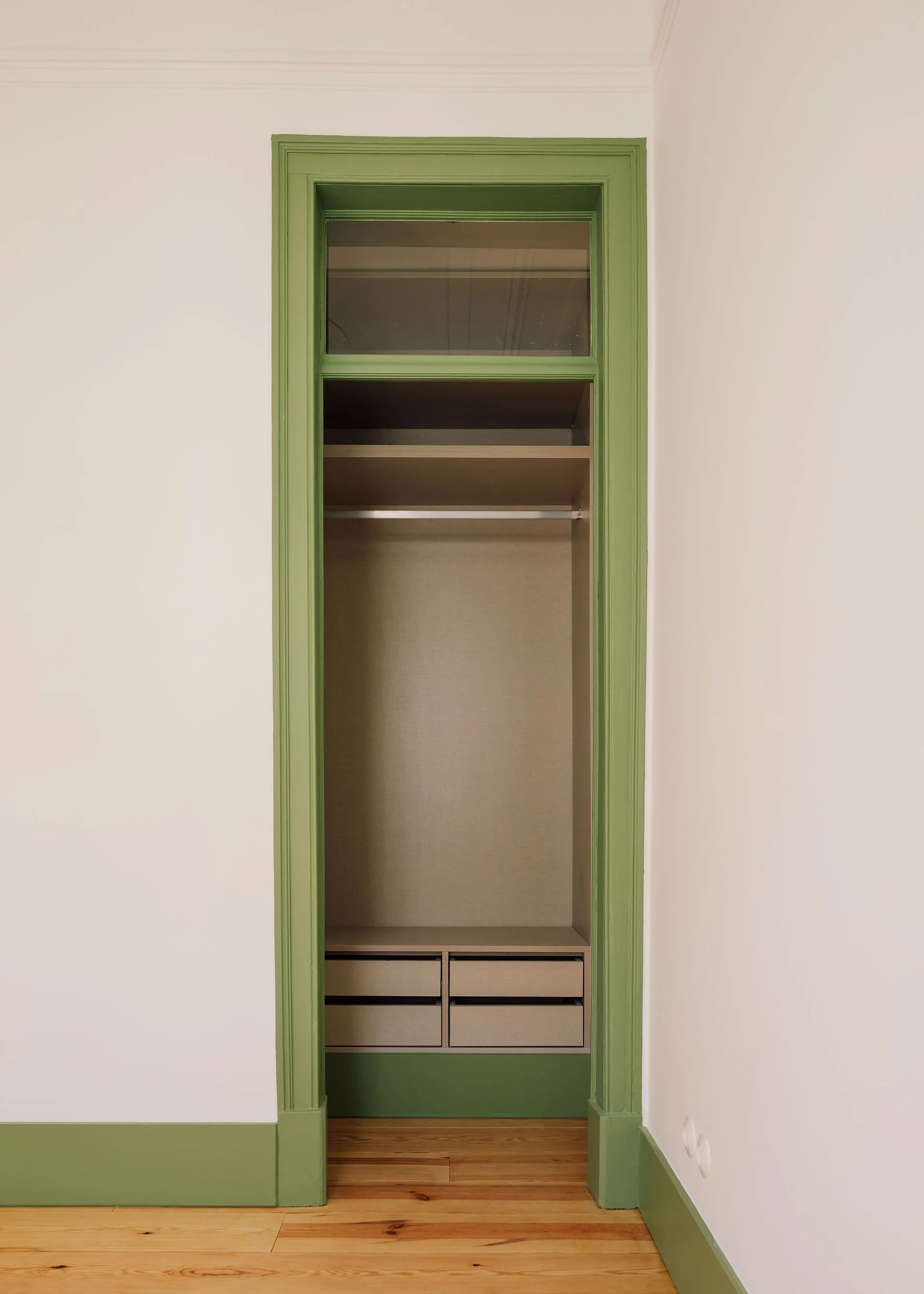 An open built-in closet with beige shelves and drawers is framed by bold green painted trim and a glass transom window.
