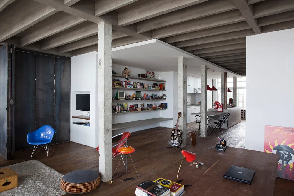 An industrial loft living room with an exposed concrete grid ceiling, concrete columns, wood flooring, and floating shelves.