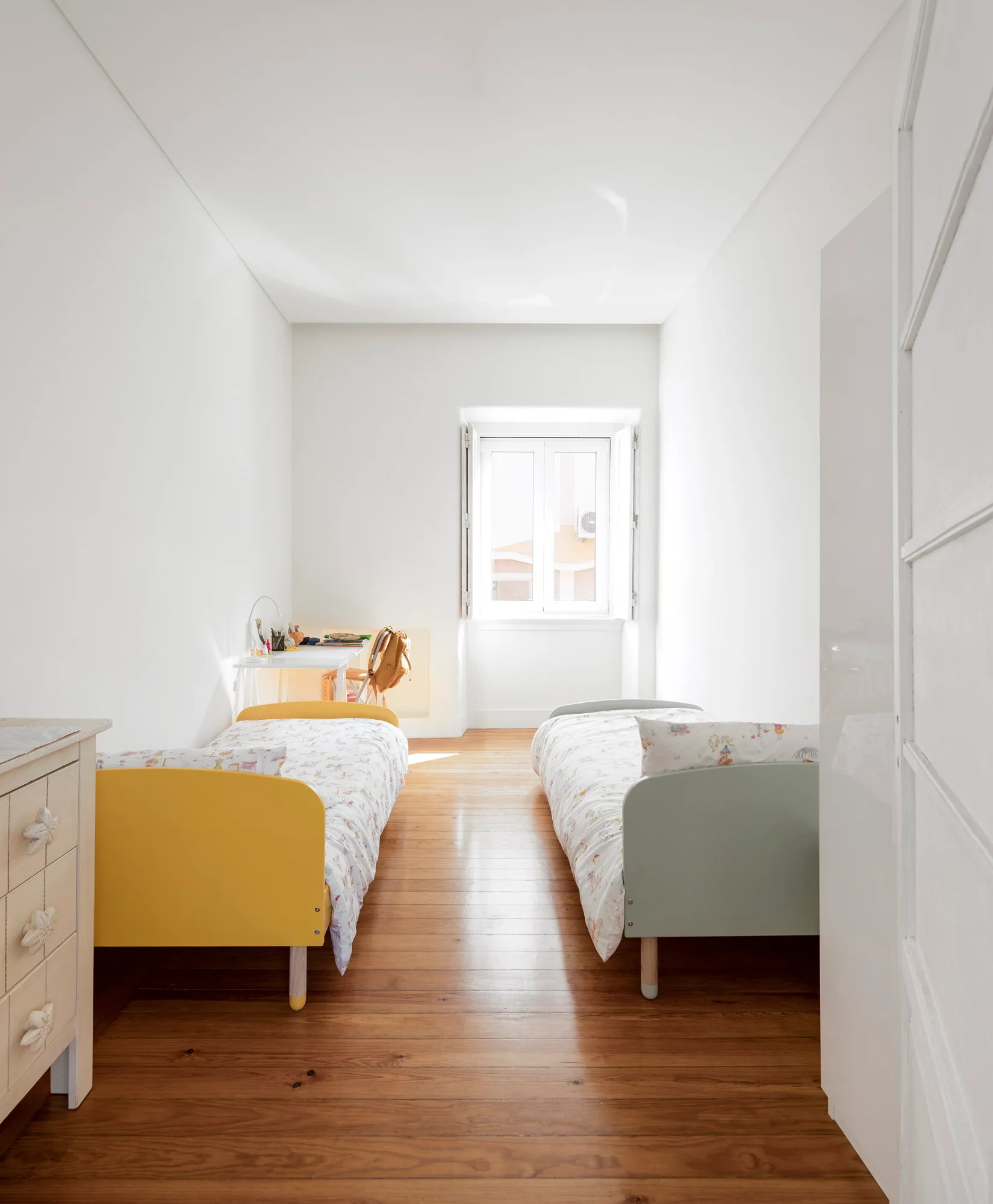 A narrow children's bedroom features two twin beds with yellow and sage green frames, wooden flooring, and a small desk near a bright window.