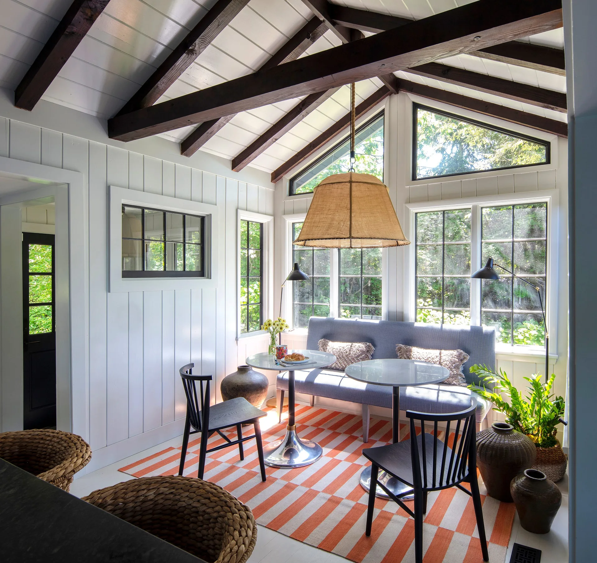 A bright dining area featuring a blue banquette, two round white pedestal tables, black chairs, and a striped orange rug under a beamed ceiling.