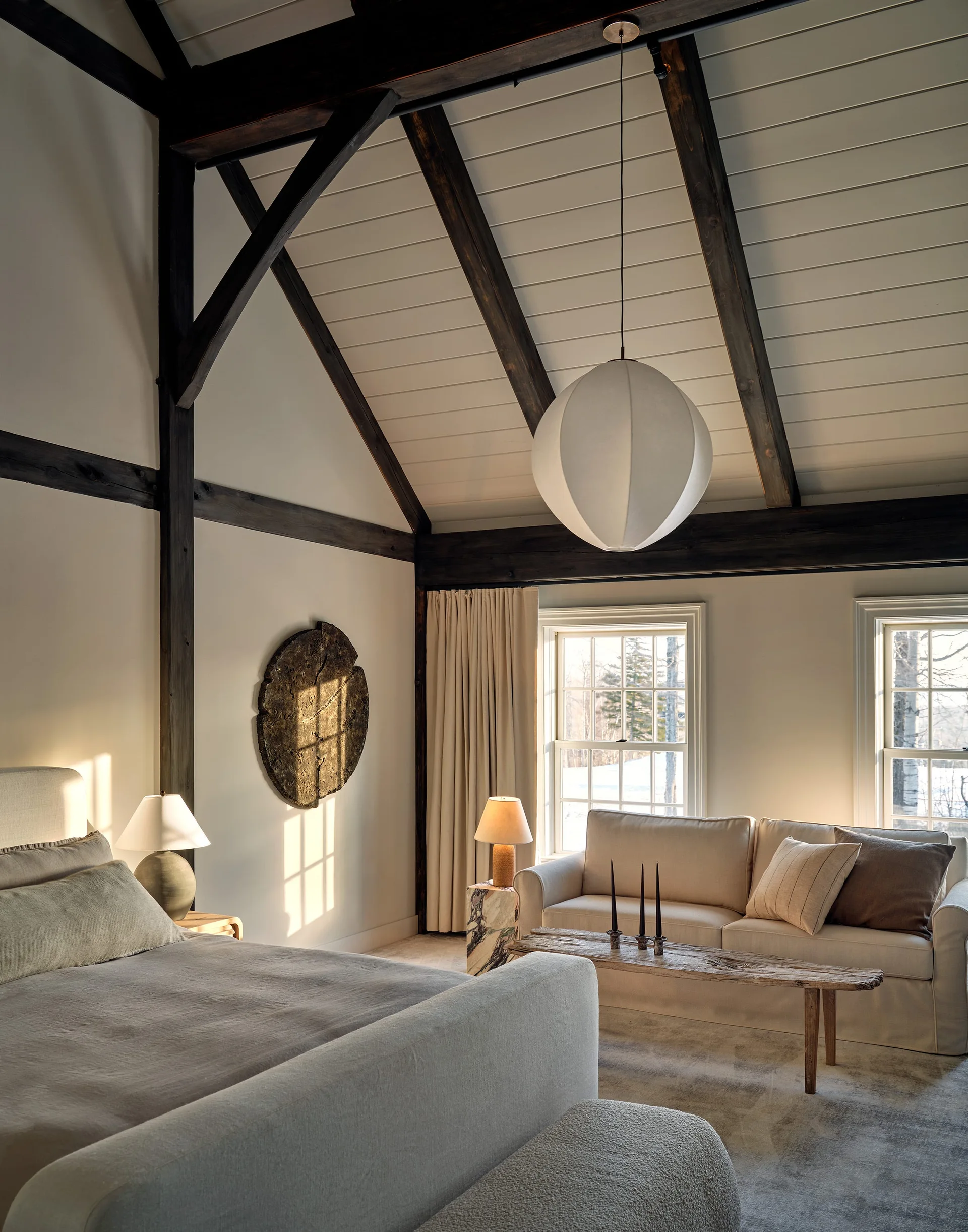A vaulted bedroom features exposed dark wood beams, an upholstered bed, a cream sofa under windows, and a large round pendant light.