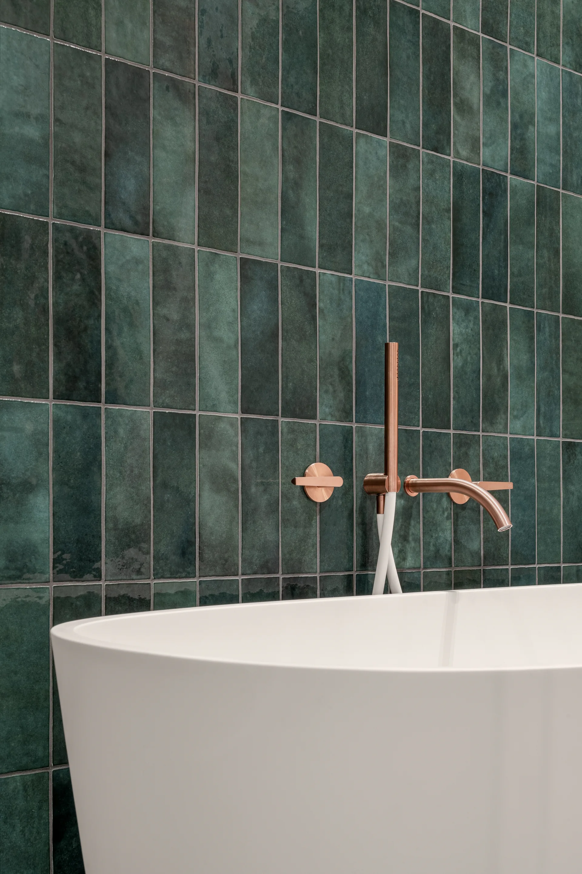 A white freestanding bathtub sits against a wall of glossy, vertically stacked green ceramic tiles featuring a wall-mounted copper faucet.