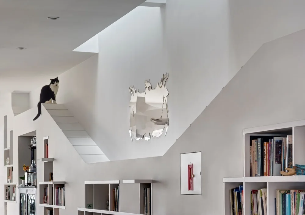 A white minimalist wall features built-in book cubbies, a liquid-shaped wall mirror, and a small step structure leading to an upper ledge.