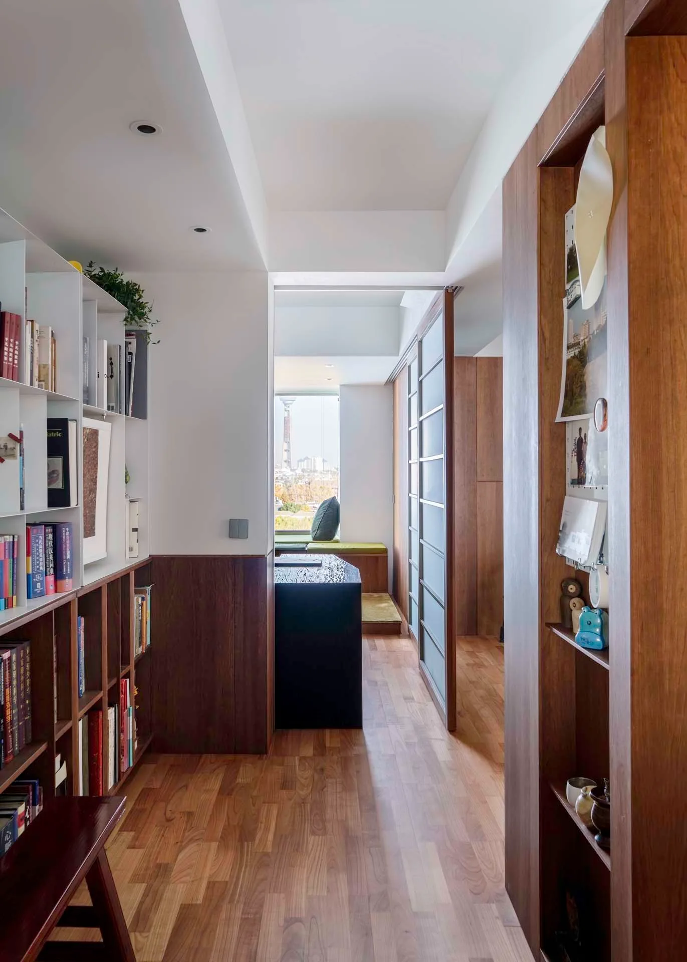 A narrow home library corridor featuring custom wood bookshelves, hardwood flooring, and a frosted glass sliding door leading to a reading nook.