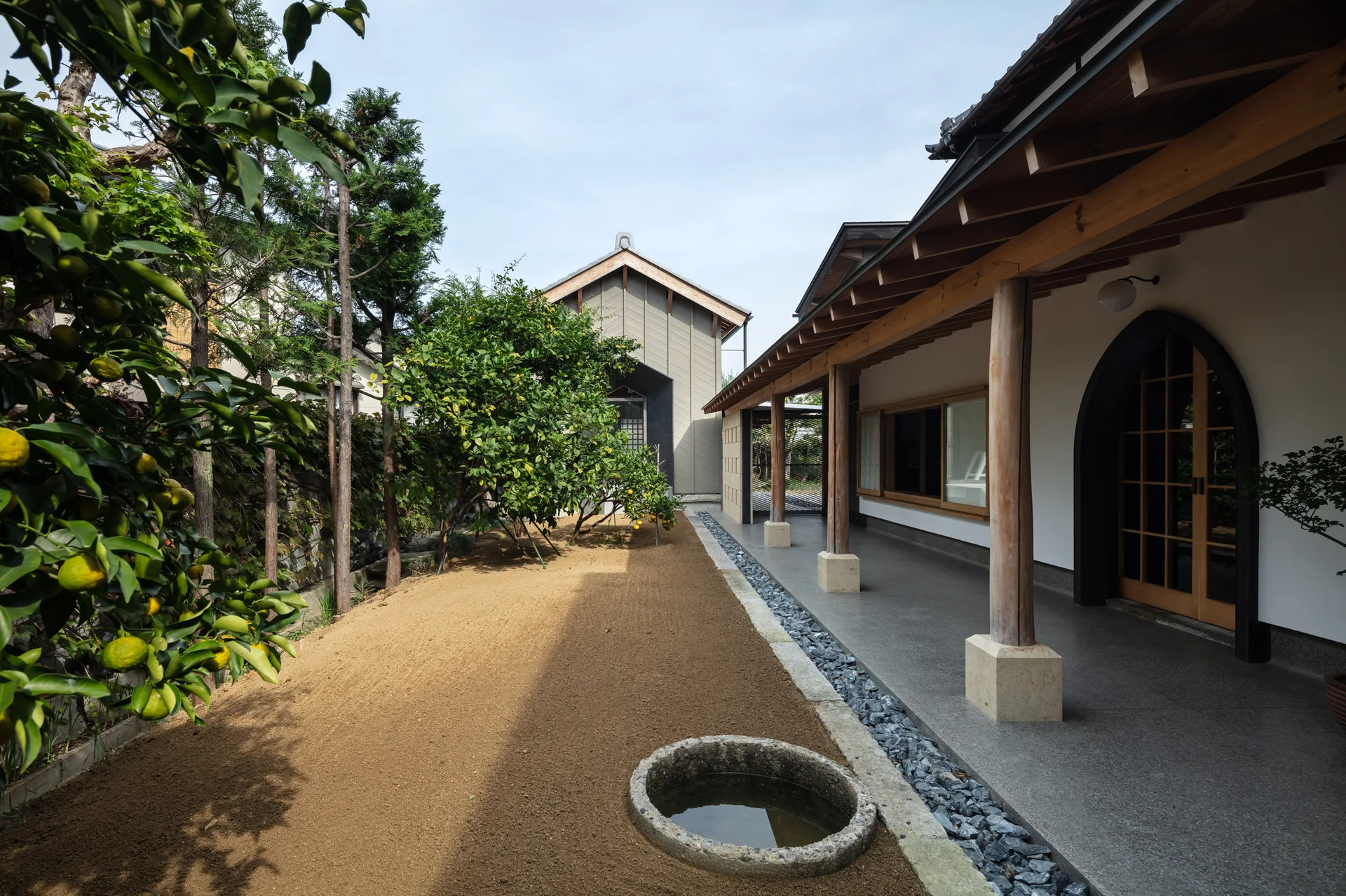 A modern Japanese house features a covered concrete porch with wooden pillars alongside a raked sand garden containing a stone water basin.