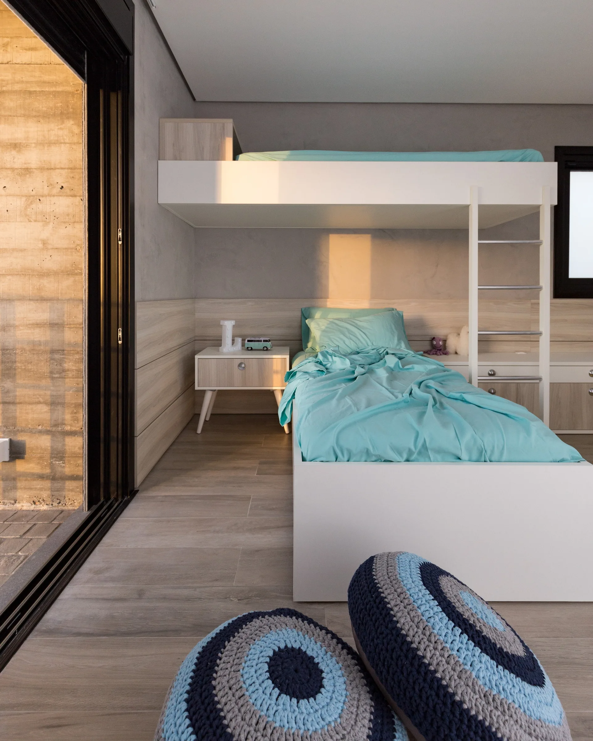 A modern kids bedroom featuring a white bunk bed with teal bedding, horizontal wood wall paneling, and blue knitted floor poufs.