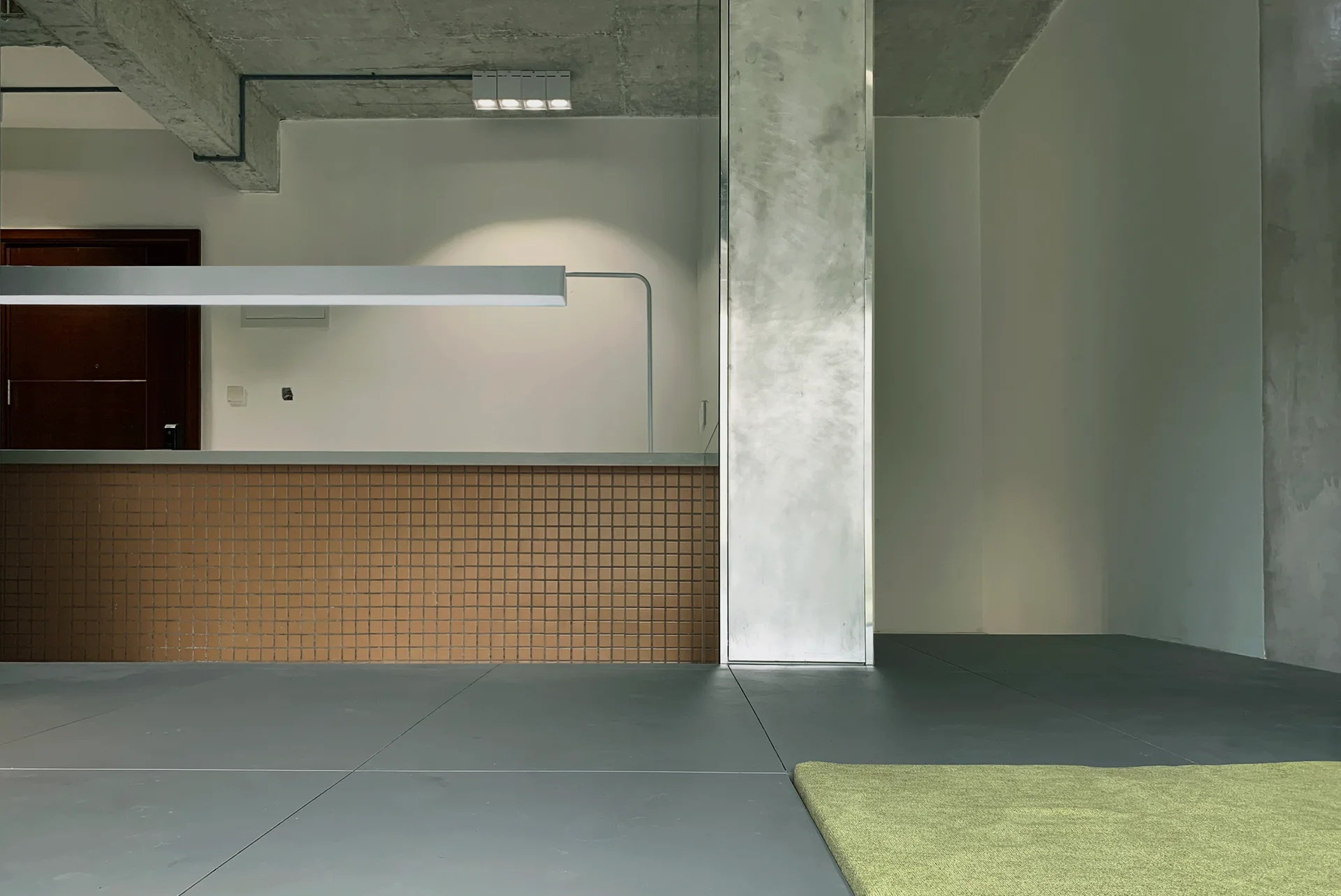 A minimalist room featuring a brown tiled counter, an exposed concrete ceiling, a large central column, and a long linear pendant light.