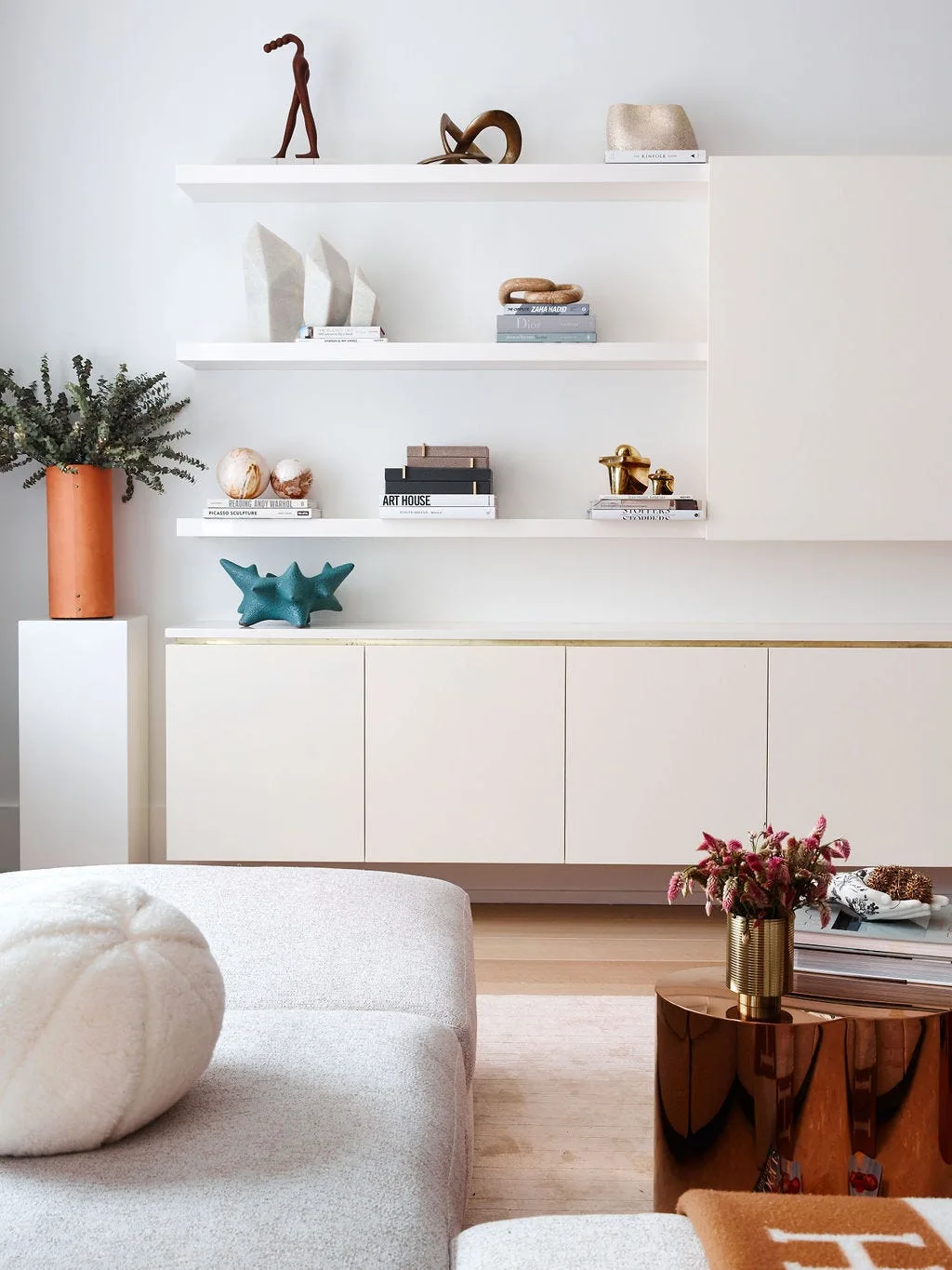 A minimalist white living room wall features built-in floating shelves and low cabinetry decorated with books and sculptures next to a copper side table.