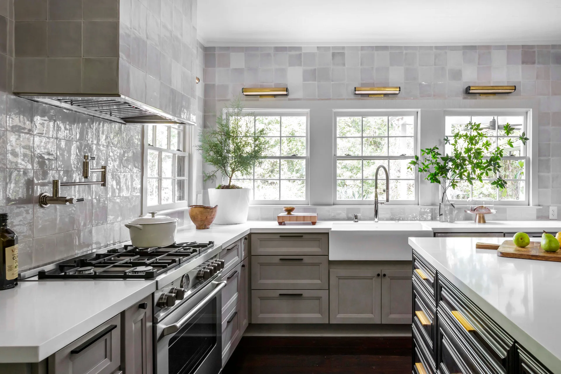 A modern transitional kitchen featuring gray cabinets, white countertops, three windows, and a glossy square tile backsplash.