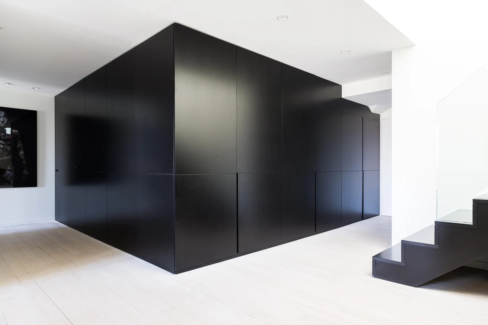 A minimalist interior corridor features a large matte black cabinetry block contrasted against light wood flooring and white walls.