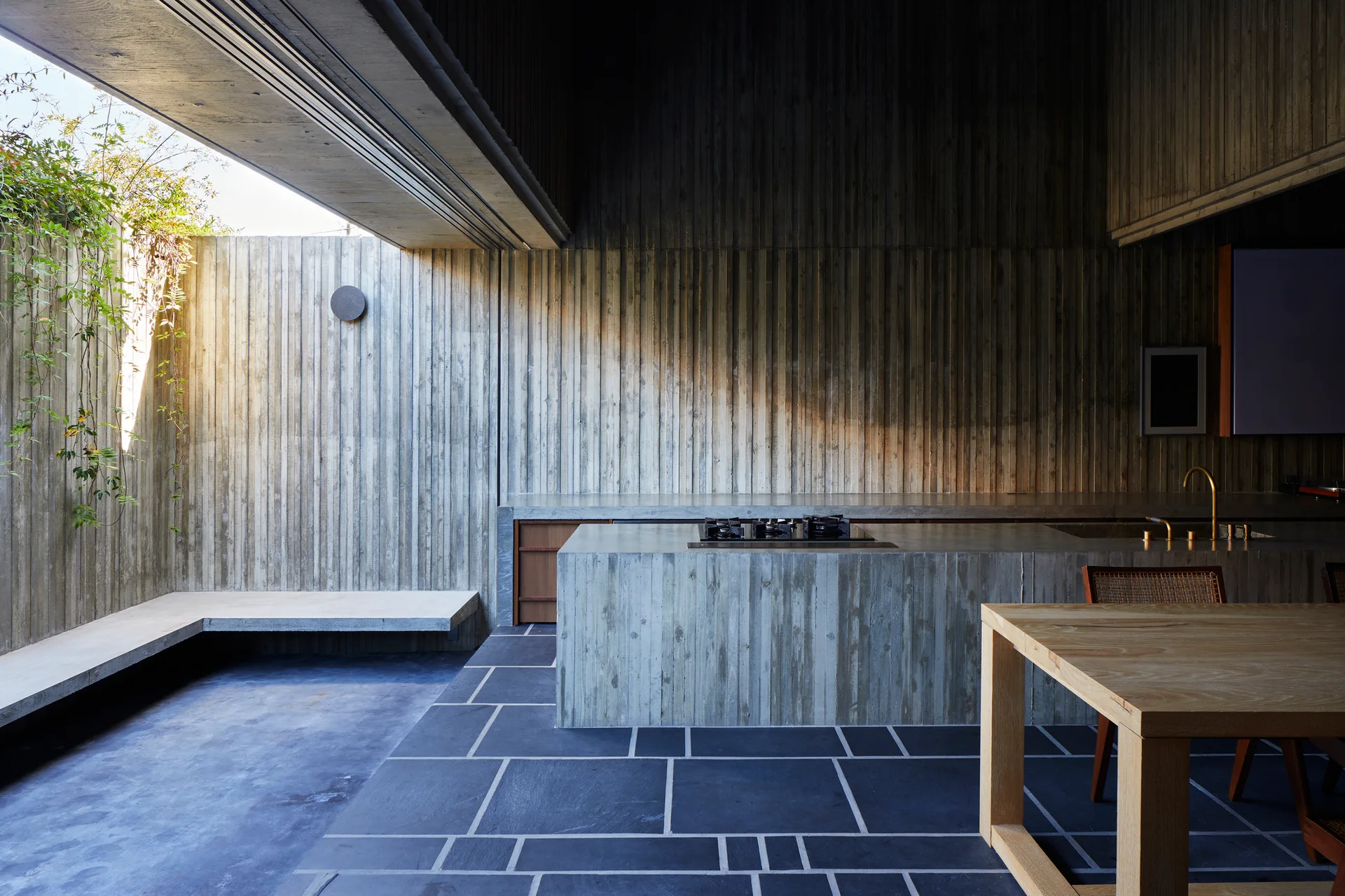 A minimalist kitchen features board-formed concrete walls and island, a dark stone tiled floor, and a wooden dining table under a skylight.