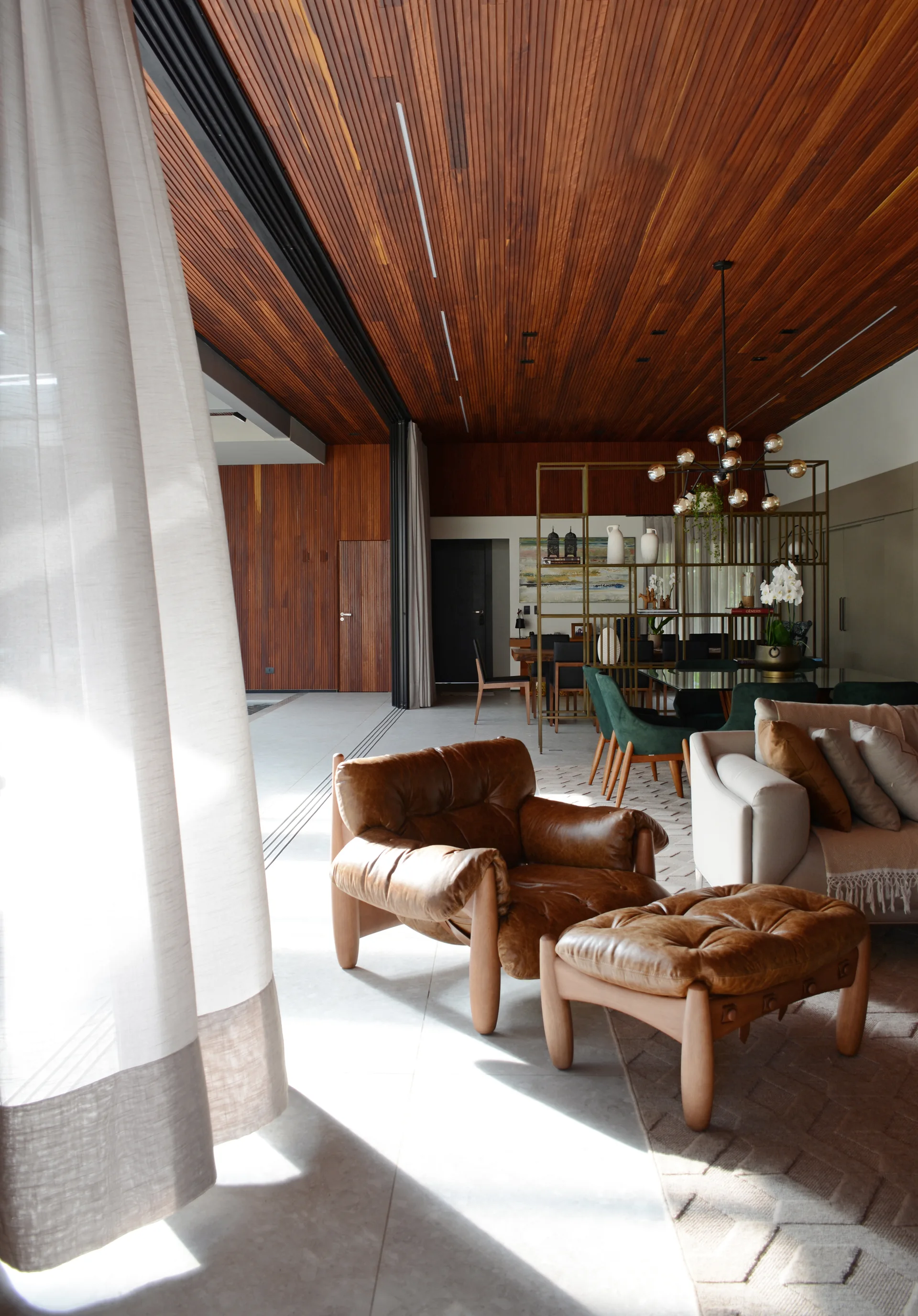 A modern open-concept living room features a tufted brown leather armchair, slatted wood ceiling, and a metal room divider.