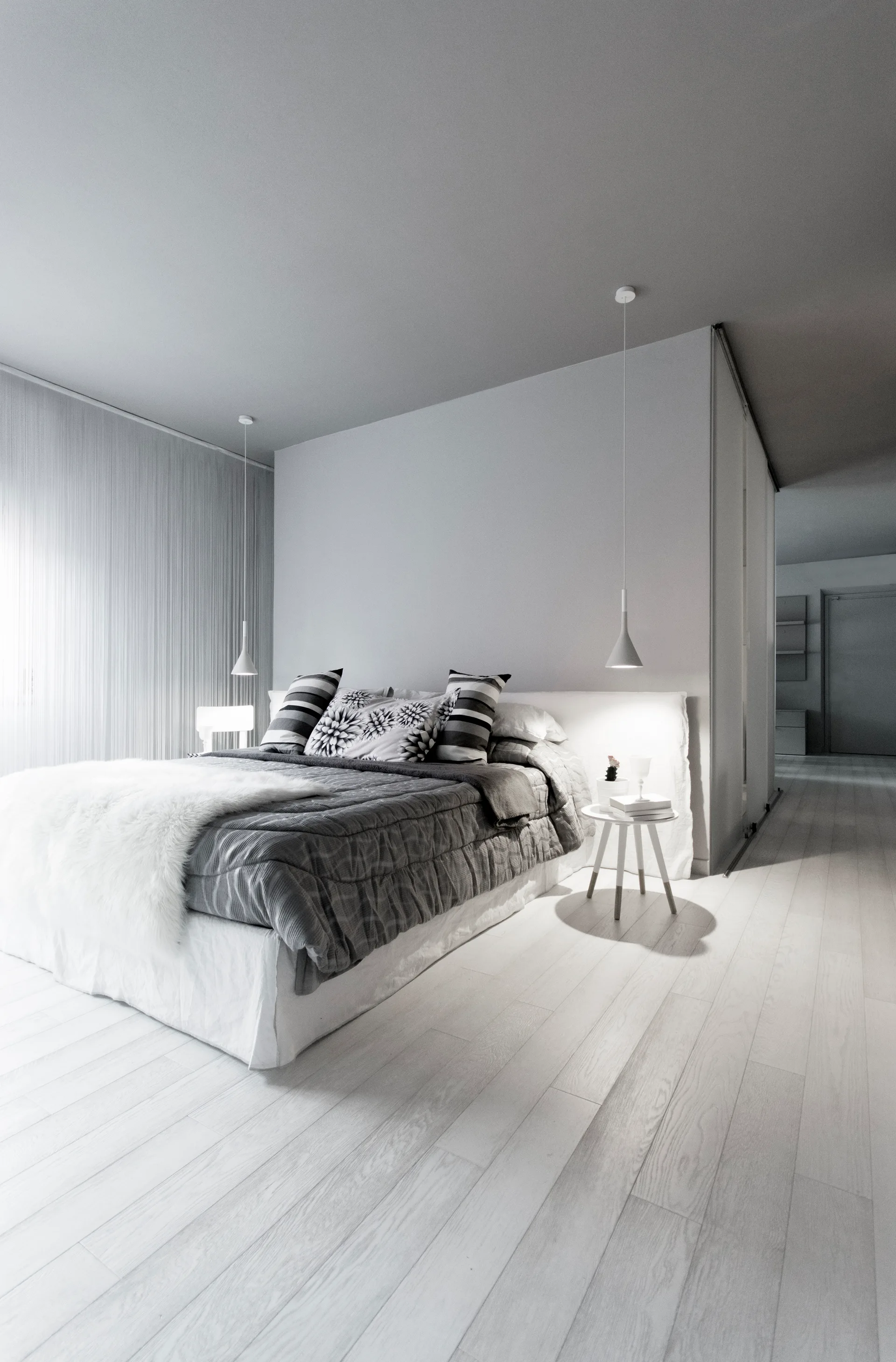 A modern bedroom features a grey bed with patterned pillows, low-hanging white pendant lights, and a white partition wall on light wood floors.