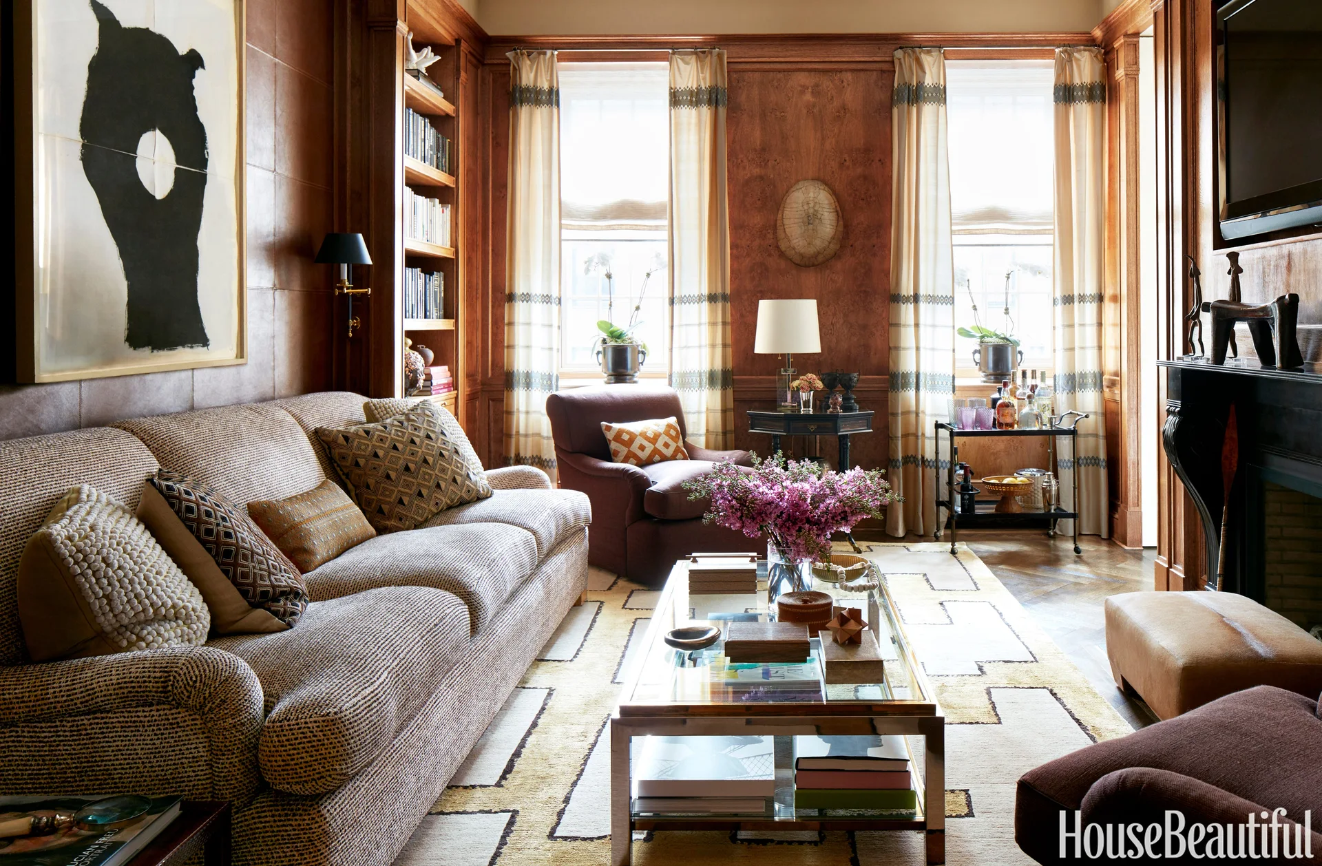 A warm wood-paneled living room features a patterned fabric sofa, a glass coffee table, and symmetrical windows with striped drapes.