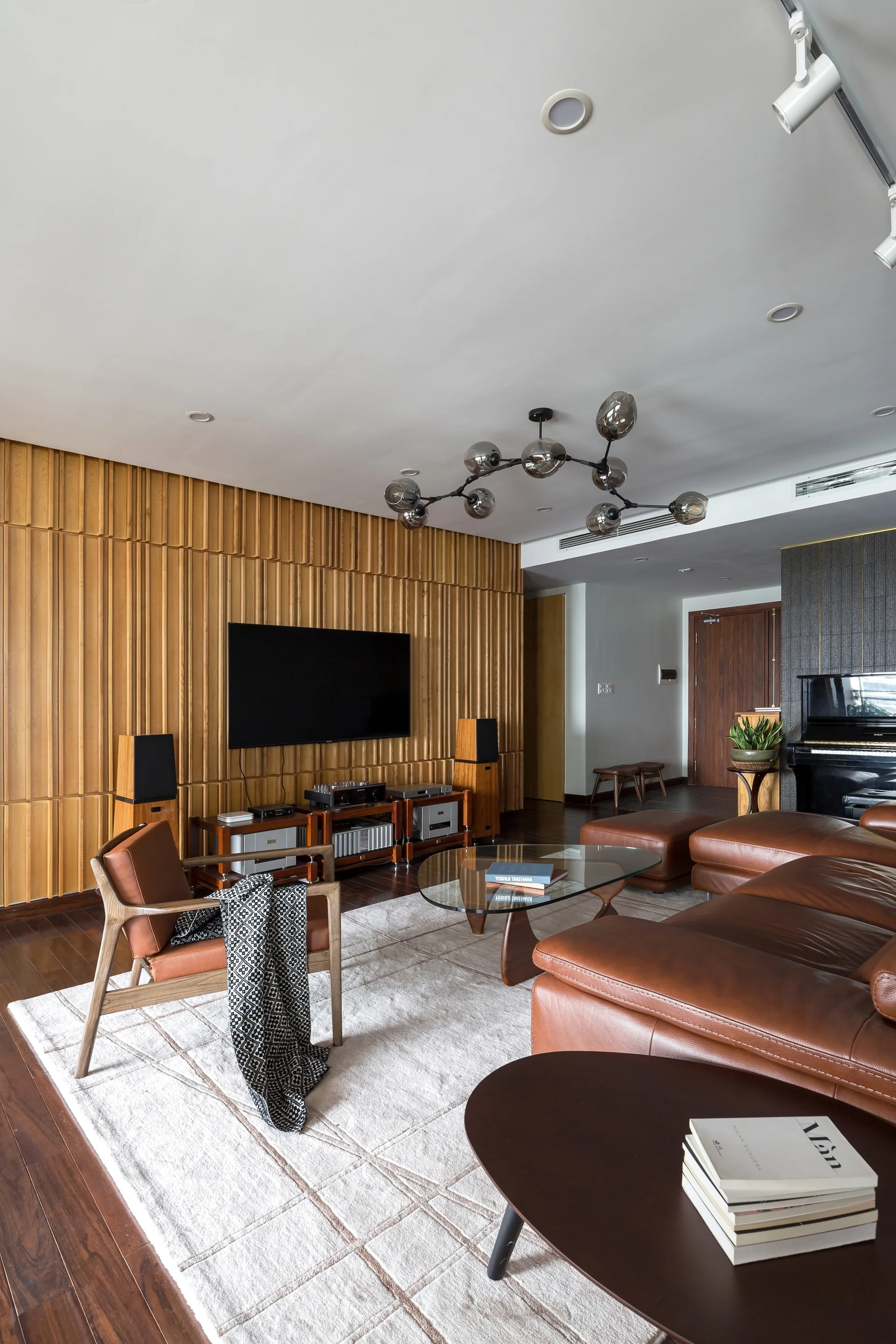 A living room featuring a slatted wood accent wall, mounted television, brown leather sectional sofa, glass coffee table, and cream area rug.