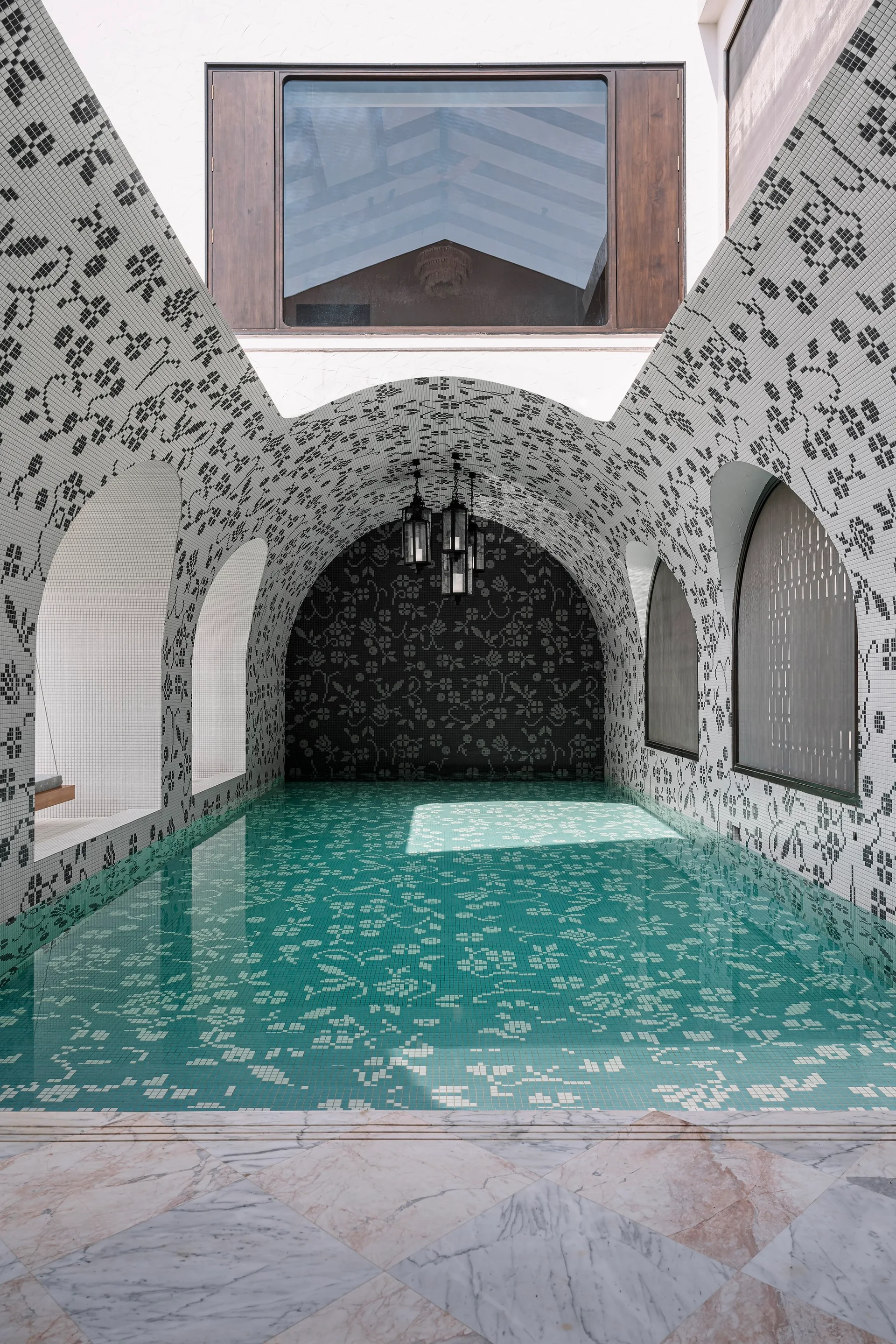 An indoor pool sits beneath a vaulted ceiling covered in intricate black and white mosaic tile patterns with arched side openings.