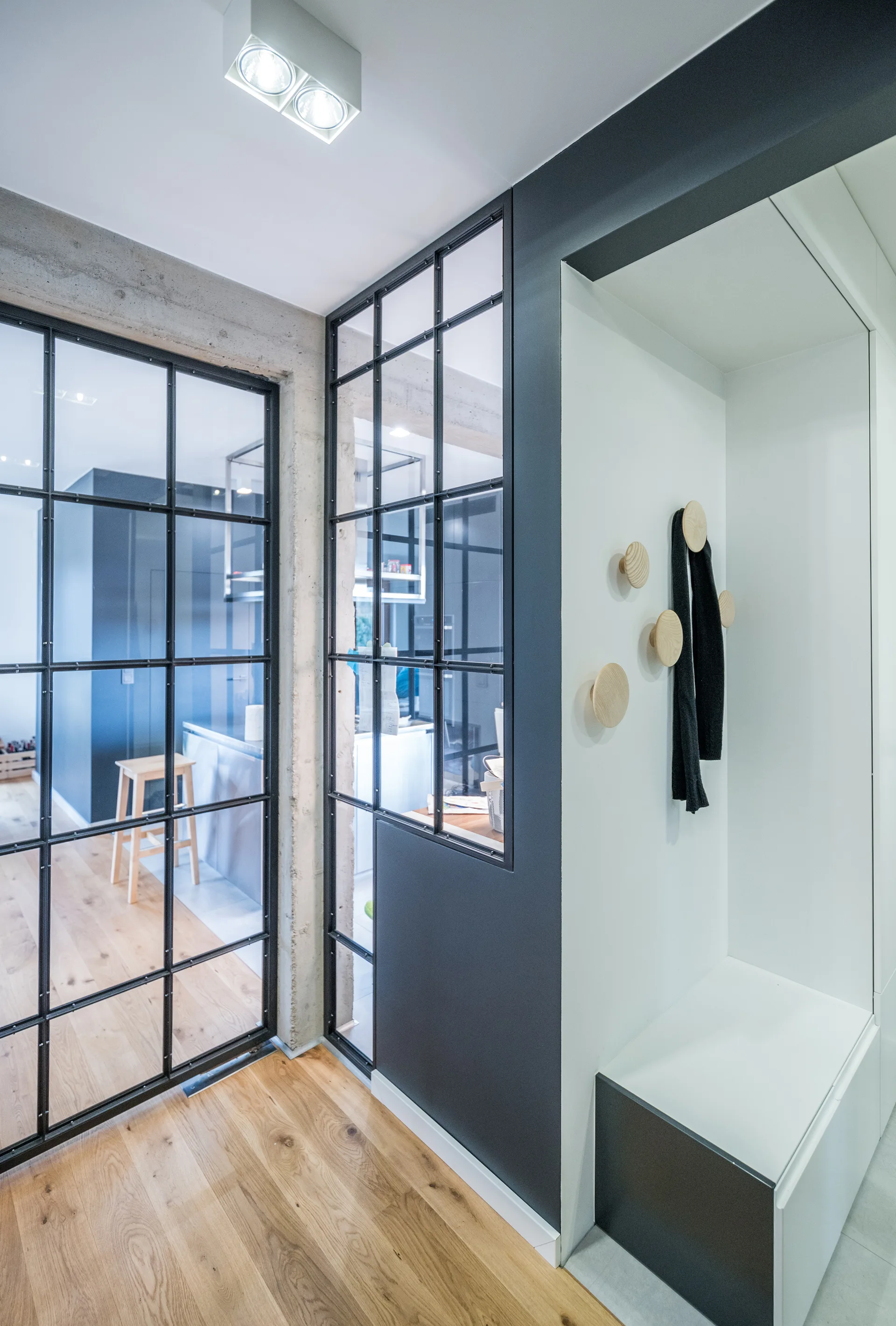 A residential entryway features a built-in white bench niche with round wooden hooks alongside a black steel-framed glass partition wall.