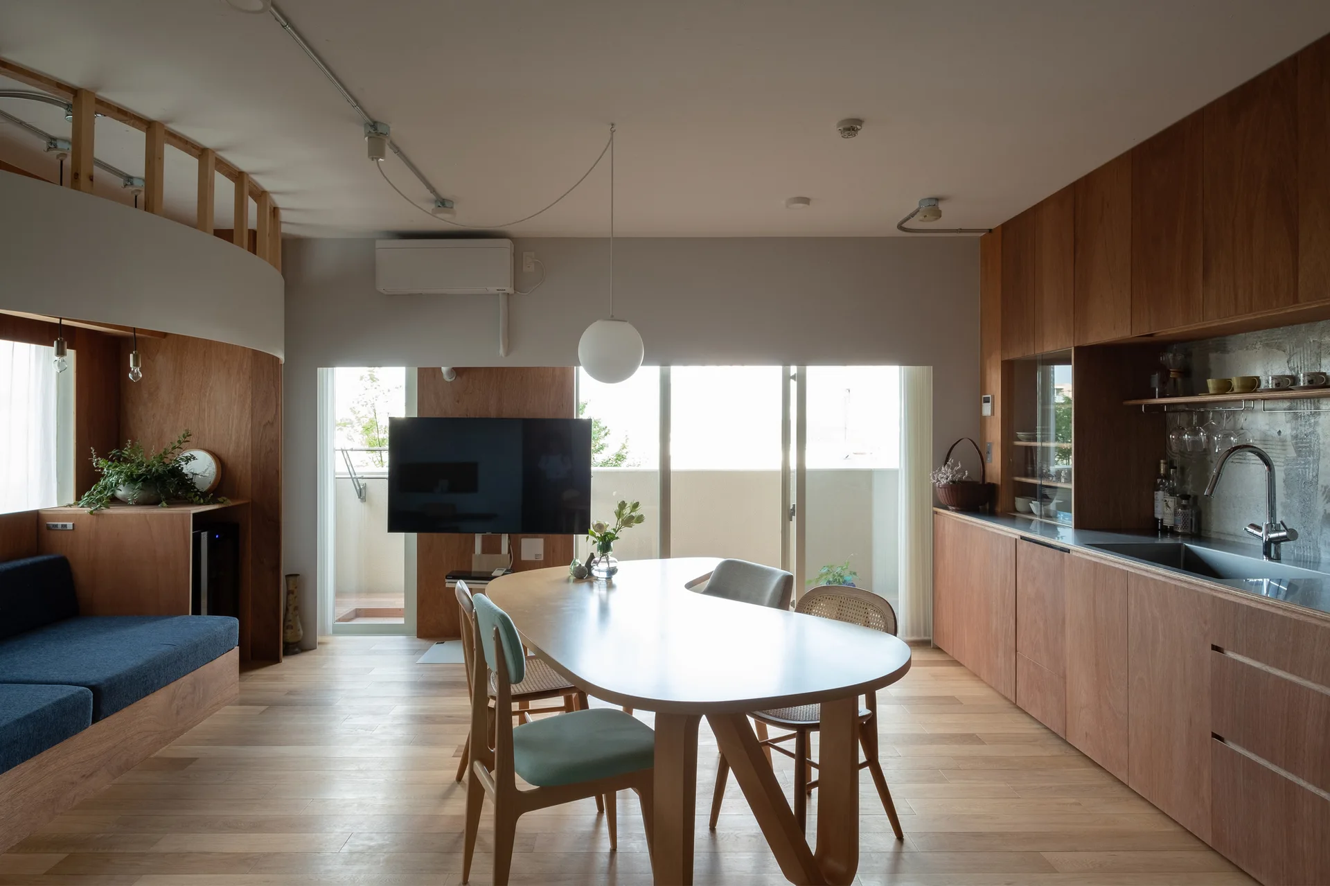 An open-plan dining room and kitchen feature warm wood cabinets, a curved wooden dining table with a globe pendant light, and a built-in blue sofa.