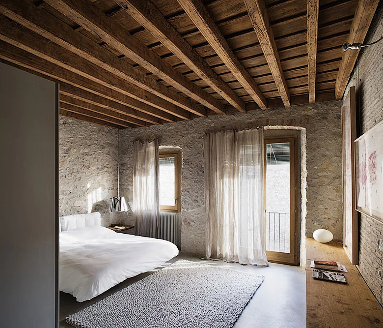 A rustic bedroom with a white bed, a grey textured rug, rough stone walls, and an exposed wooden beam ceiling.