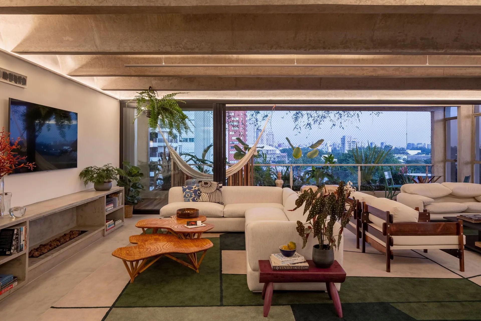 A modern living room features a beige sectional sofa, organic wood coffee tables, and exposed concrete ceiling beams next to a balcony with a hammock.