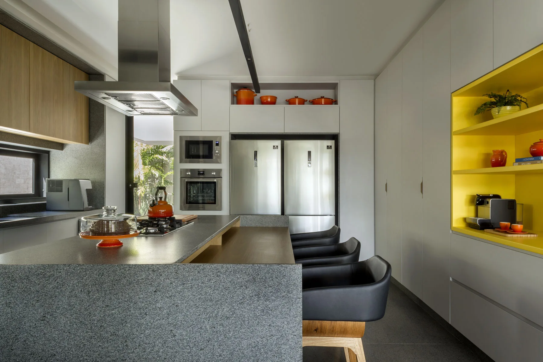 A modern kitchen features a grey stone island with black bar stools, flat-front white cabinets, and a built-in bright yellow shelving niche.