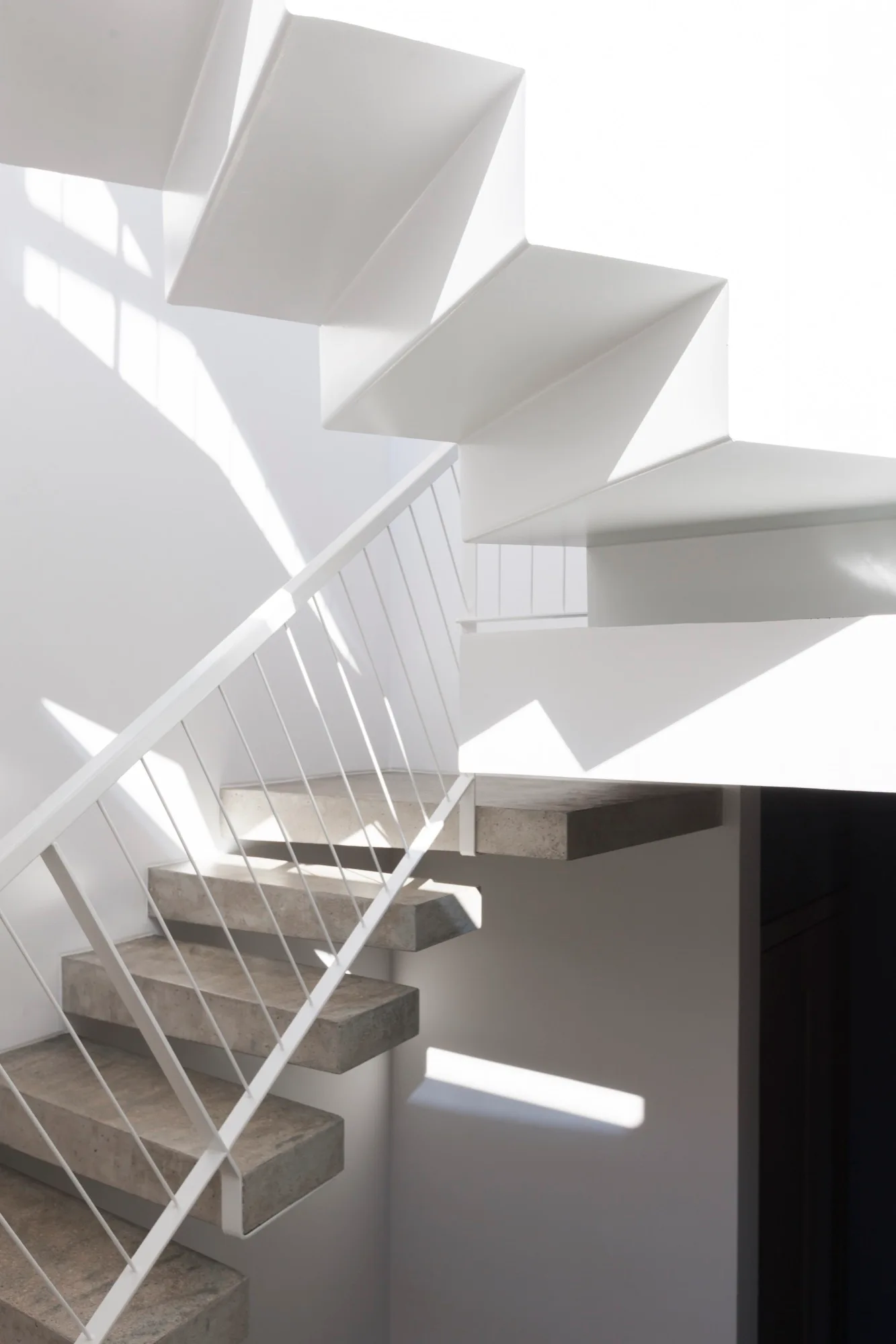 A modern staircase featuring floating concrete treads and a thin white metal railing with sharp shadows cast on a white wall.