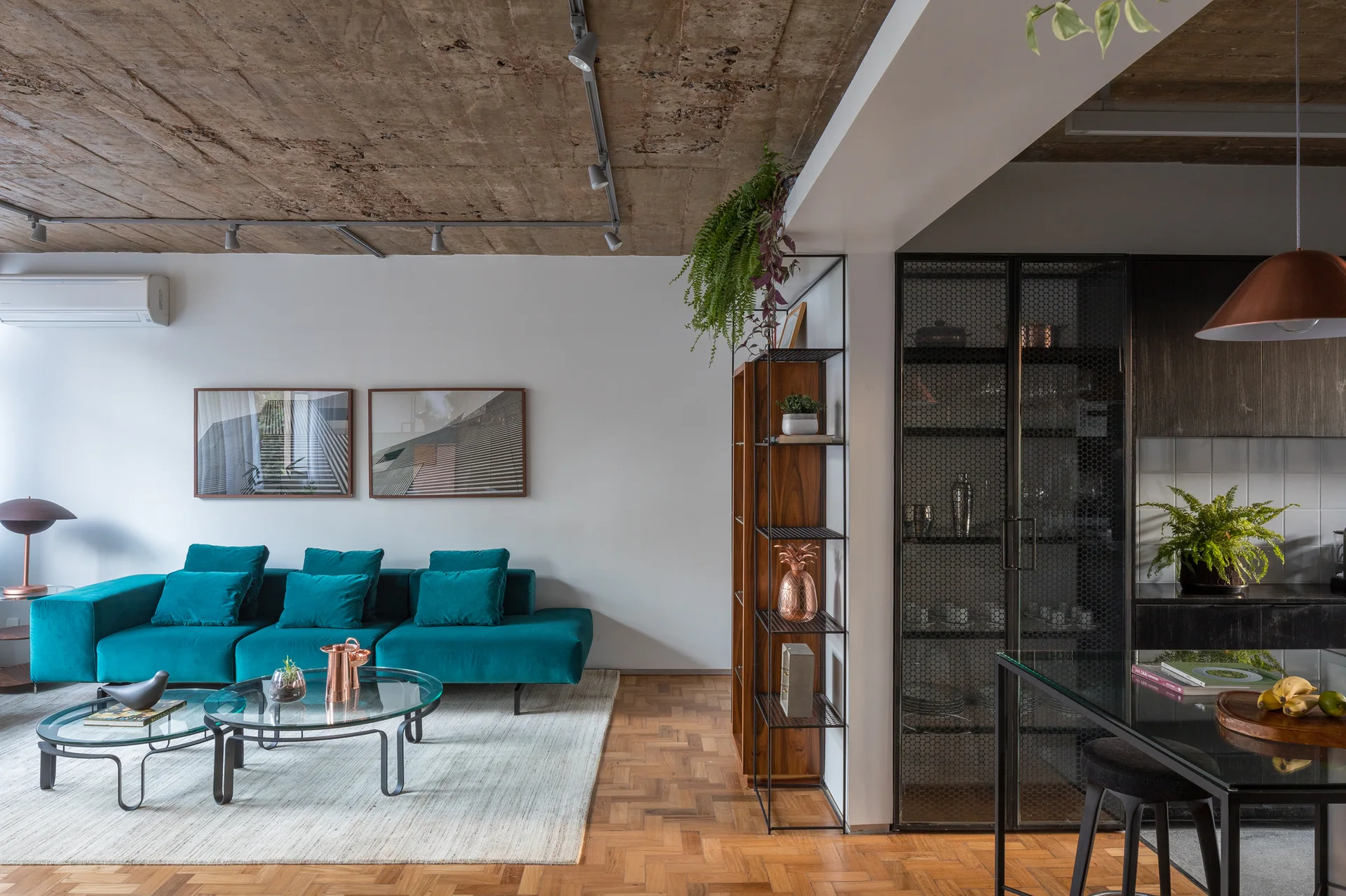 An open-plan living room features a bright teal velvet sofa on a light rug, under an exposed concrete ceiling with warm wood parquet flooring.